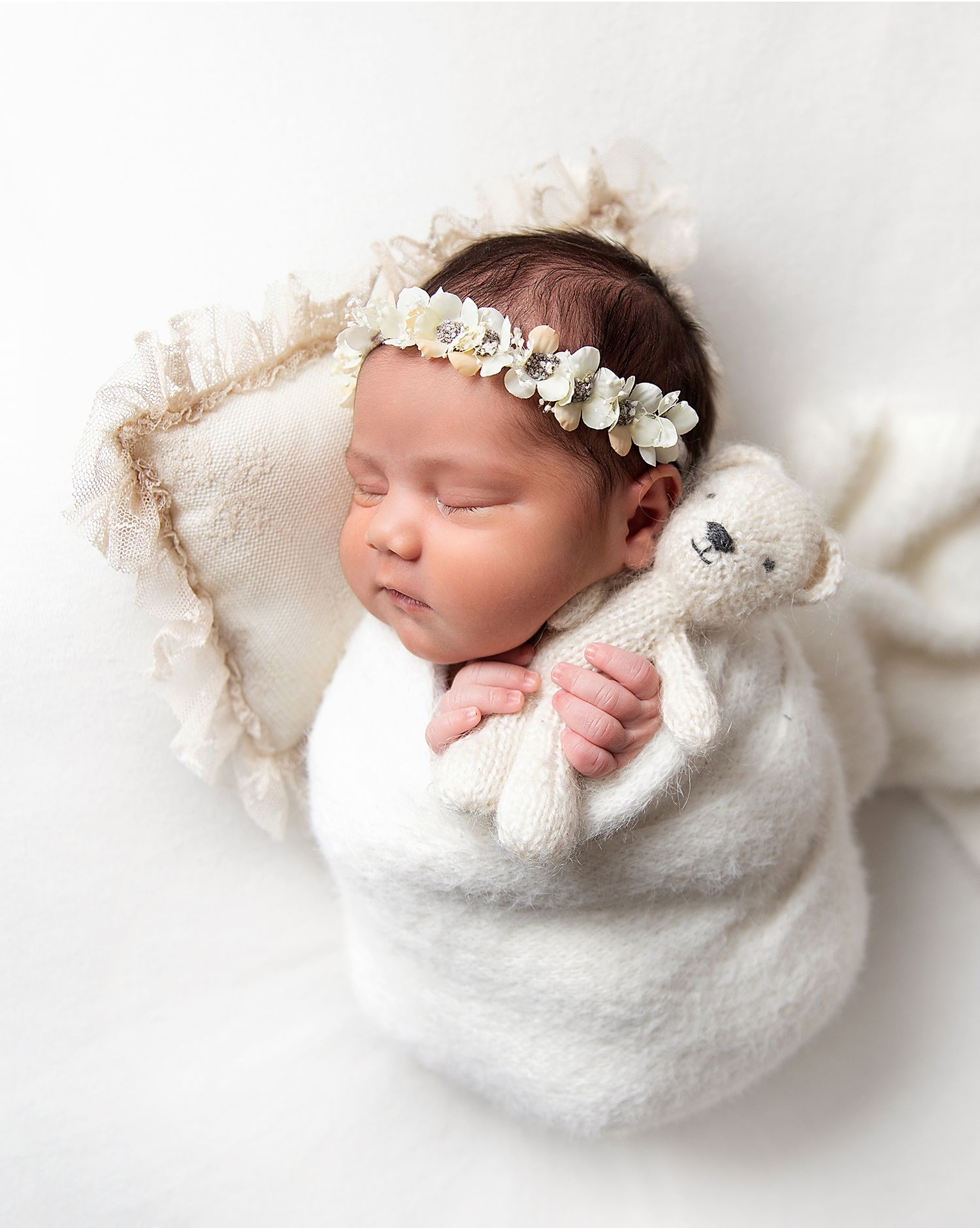 Newborn baby sleeping peacefully wrapped in soft white blanket with delicate floral crown headpiece. During a newborn photoshoot in Bexley