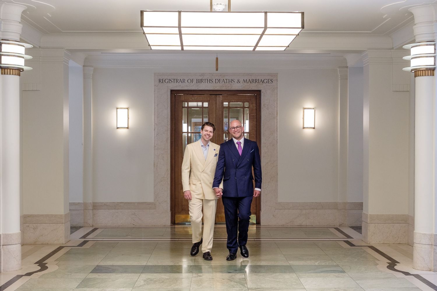 Two people in formal attire walk together through an elegant hallway with white walls and sconce lighting.