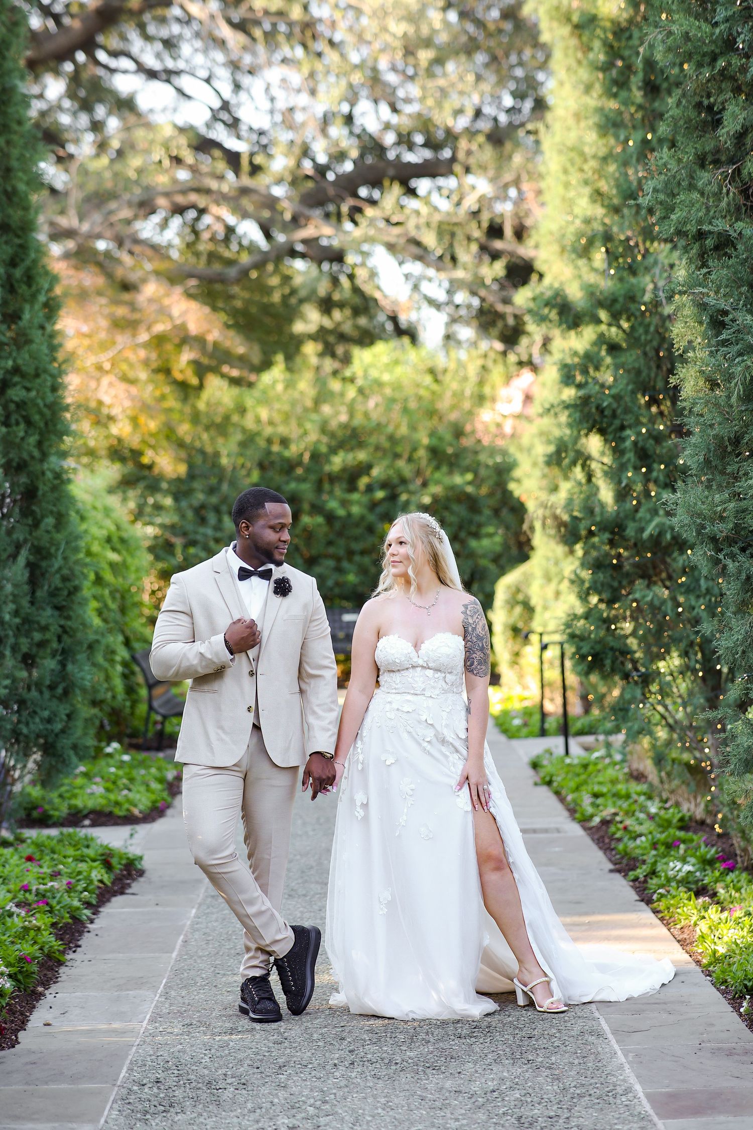 A romantic wedding couple poses together on a garden pathway with by lush greenery and flowers on their special day at the Dallas Arboretum.
