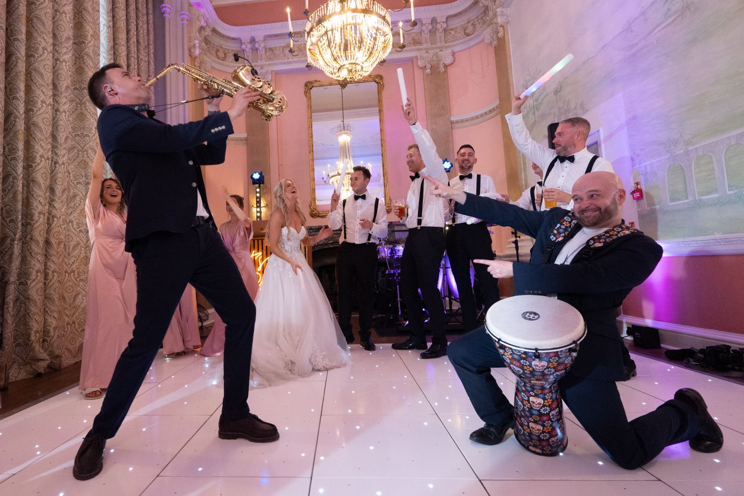 Wedding photographers capture a fun moment of musicians and guests dancing in an elegant ballroom with chandeliers.