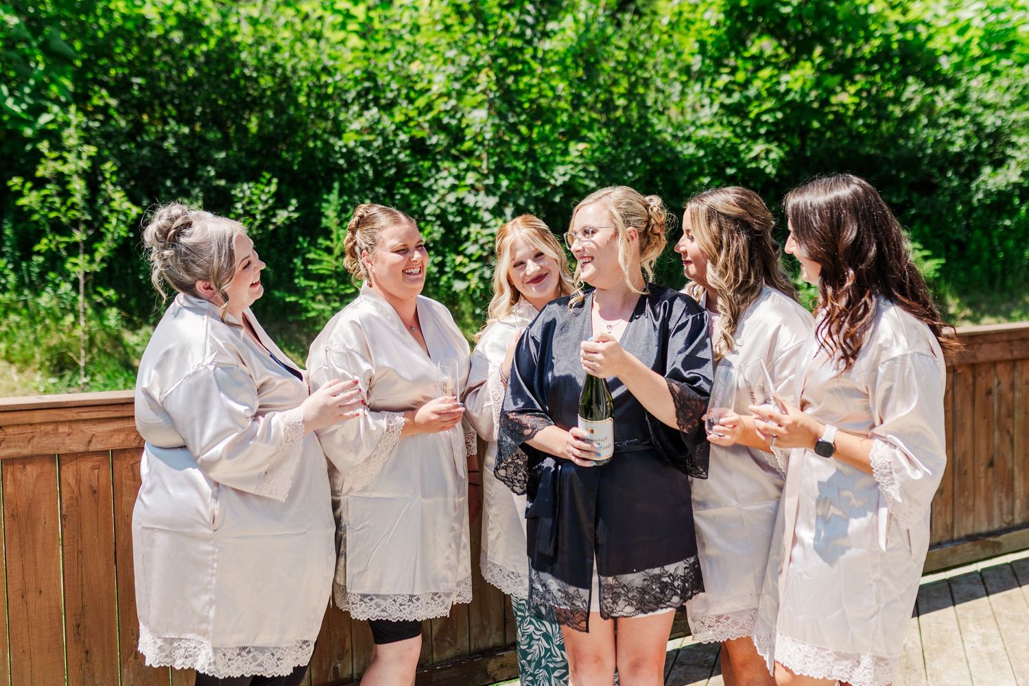 A group of women in white and black robes share laughs and drinks on a wooden deck surrounded by green foliage.