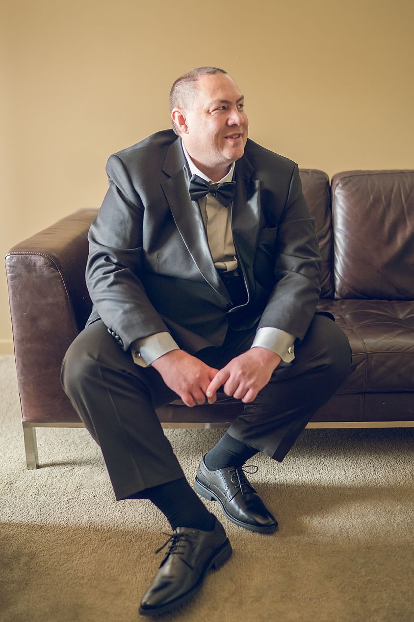 Someone wearing a dark suit and bow tie sits on a brown leather couch looking to the side in formal attire.