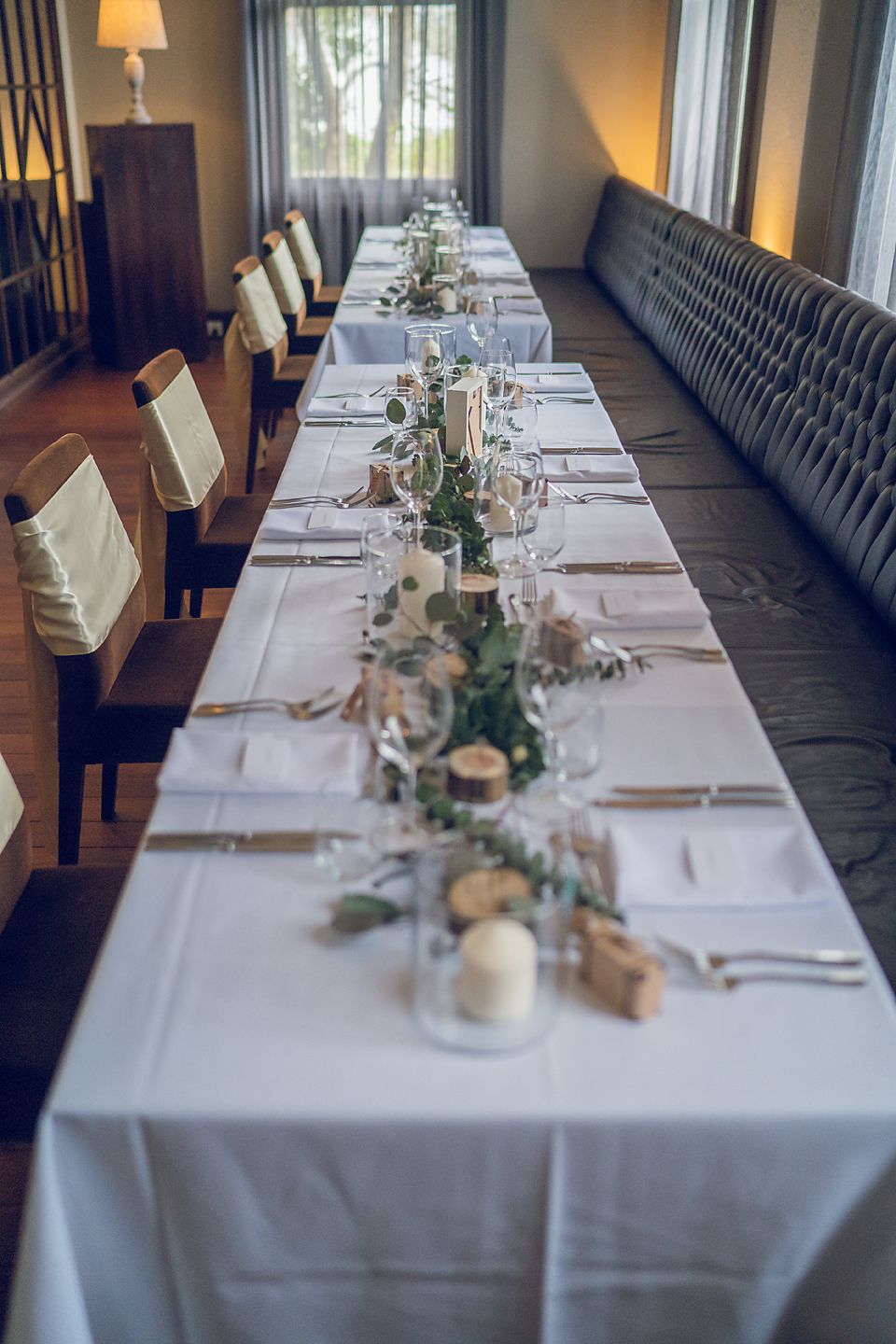 Elegant long dining table set with white linens, rustic centerpieces and candles alongside a tufted grey banquette.
