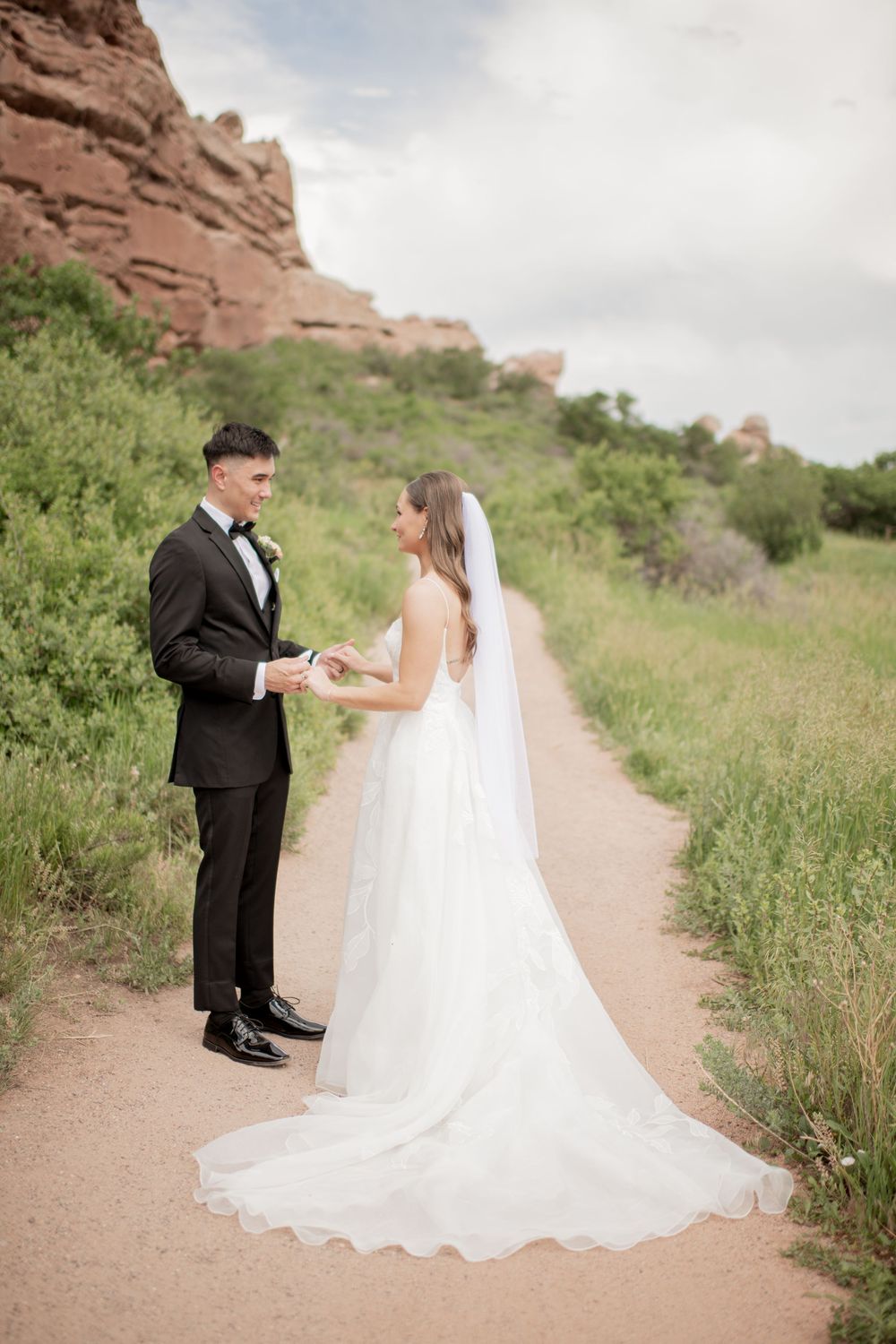 A bride and groom share a romantic moment on a dirt path with red rock formations and green foliage in the background.
