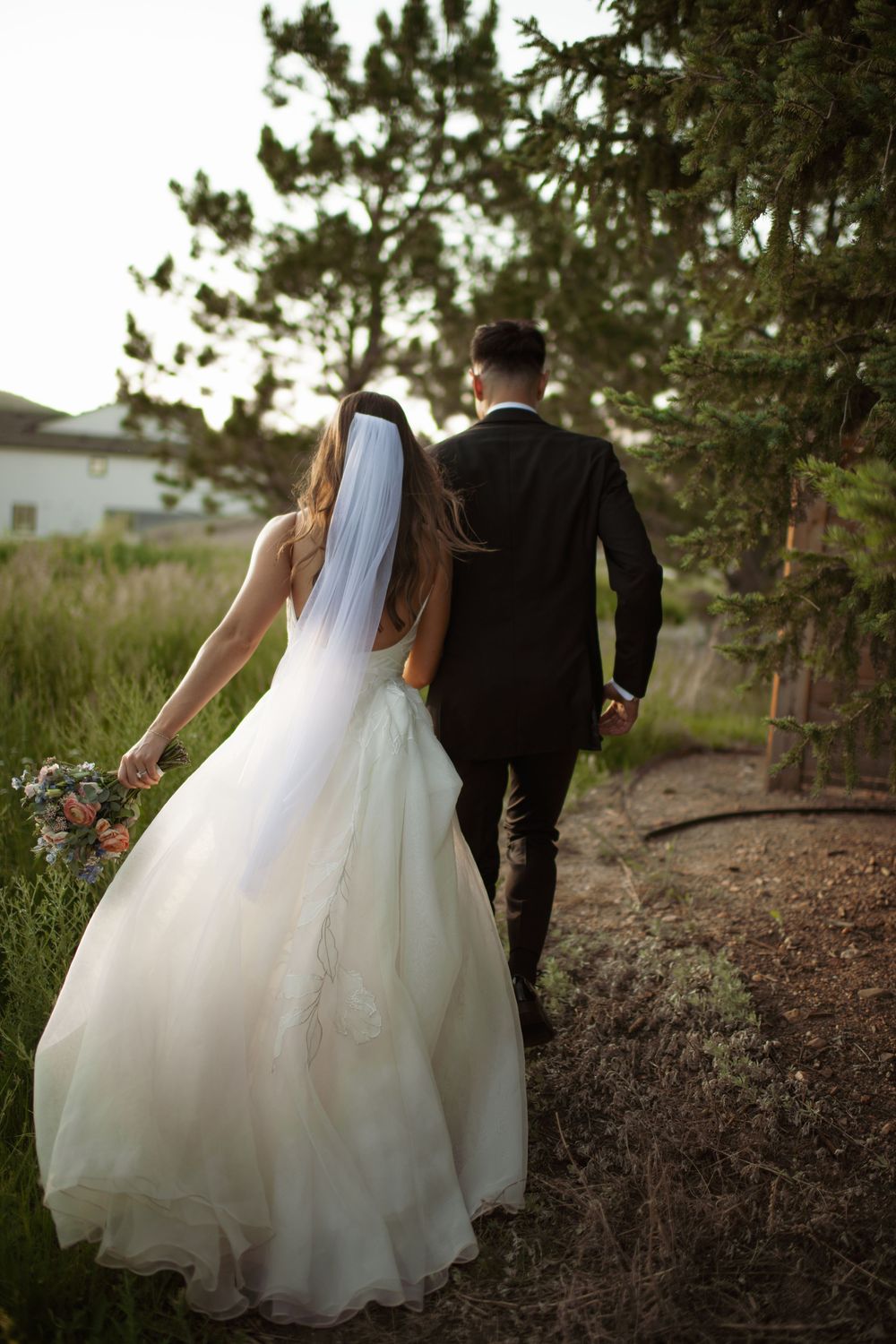 A bride in a flowing white gown and groom in black suit walk together on a rustic path surrounded by evergreen trees.