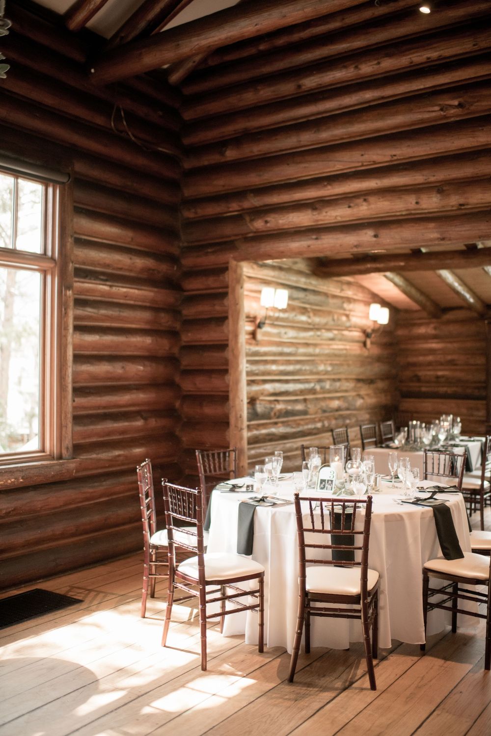 Rustic log cabin wedding venue interior with wooden walls and round tables set with white linens and chiavari chairs.