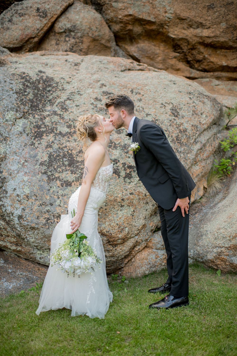 A couple shares a romantic kiss against a natural rock face backdrop at an outdoor wedding ceremony.