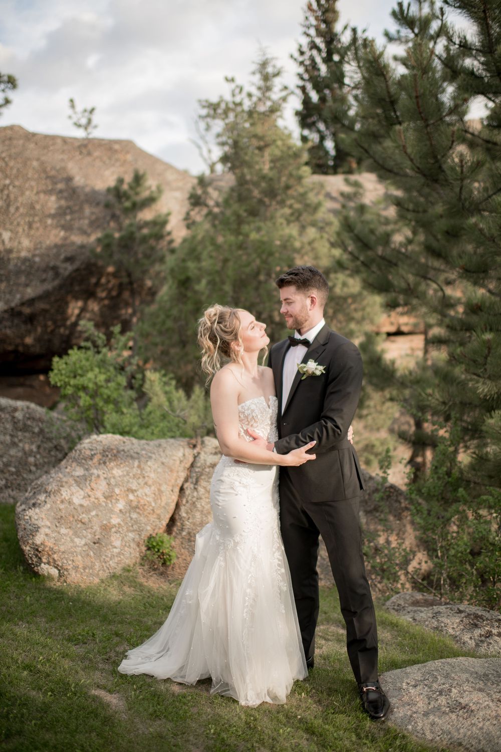 A romantic wedding couple poses in front of large boulders and pine trees in a scenic mountain setting.