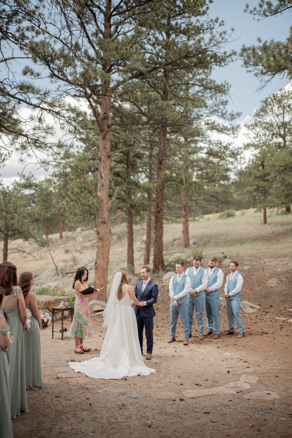 An outdoor wedding ceremony takes place in a scenic mountain setting surrounded by tall pine trees on a sloping hillside.
