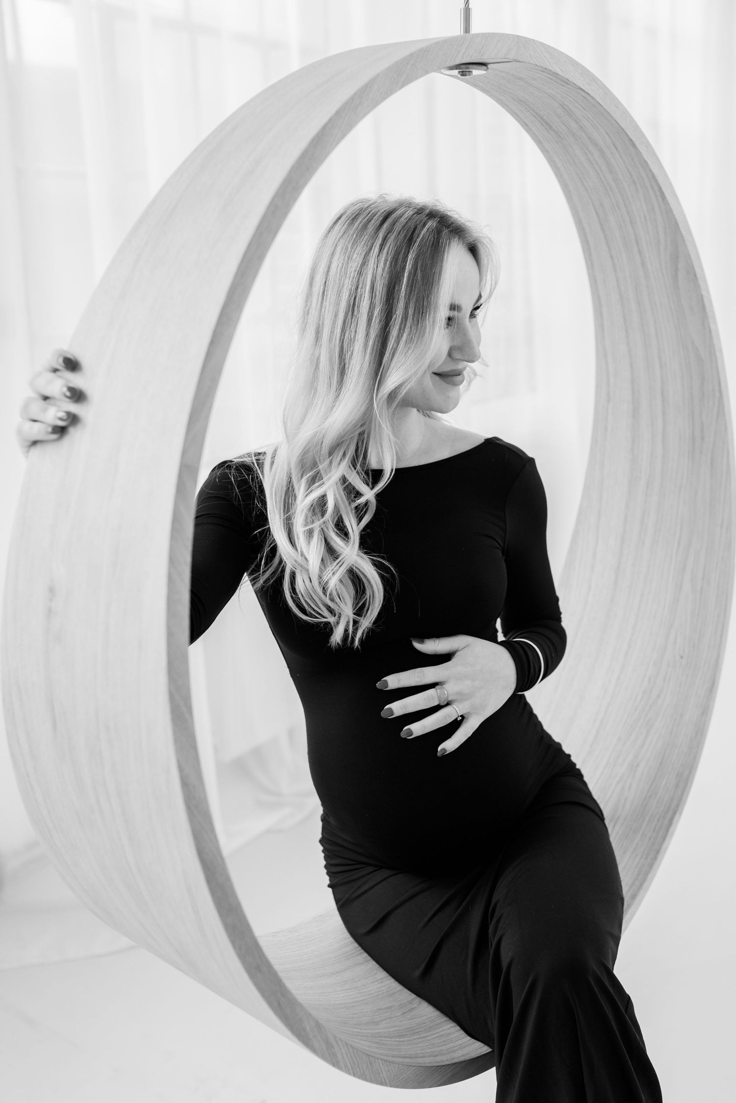 Black and white maternity photo of woman in black dress sitting in circular window frame.