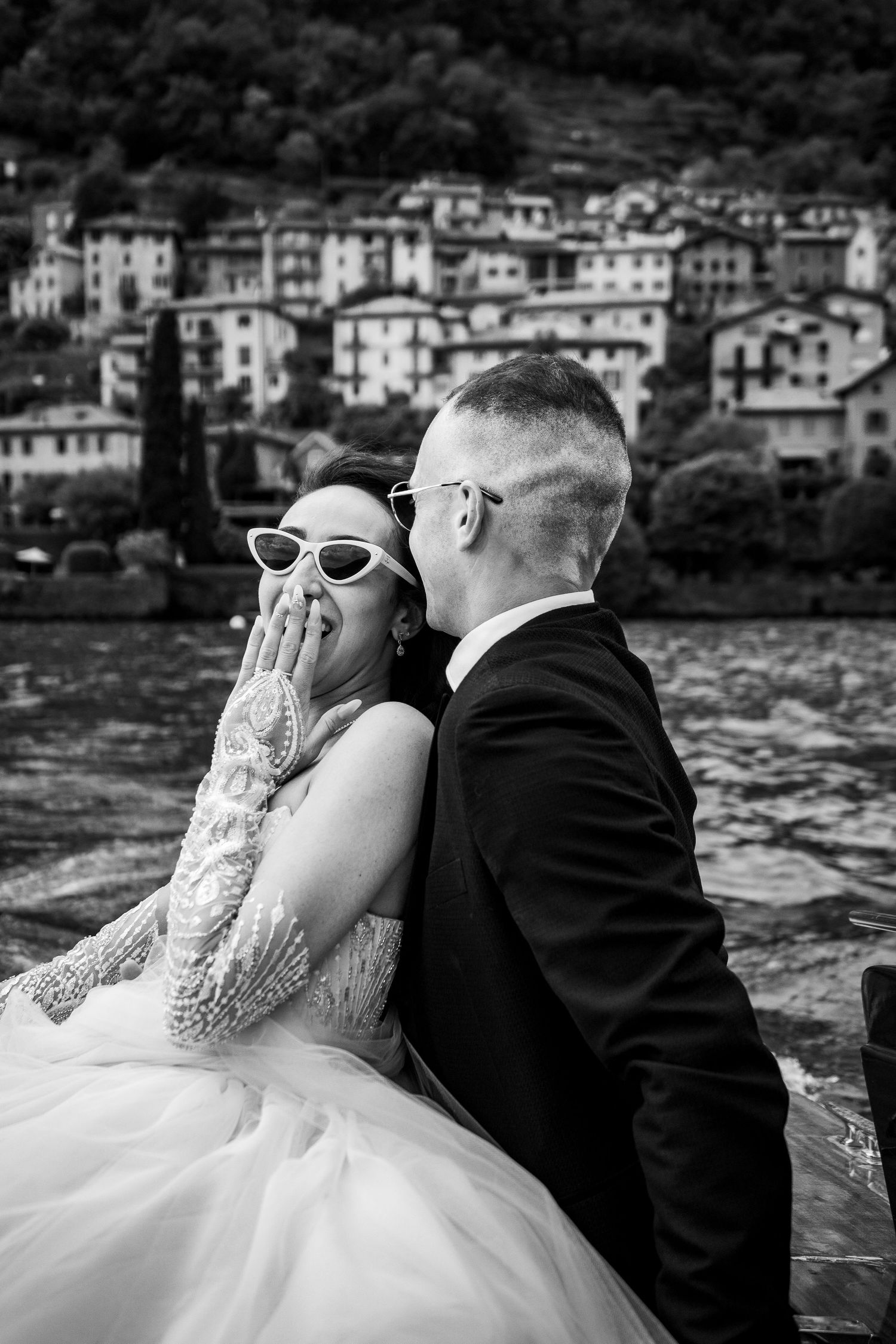 Black and white wedding portraits of couple by waterfront with scenic village backdrop.
