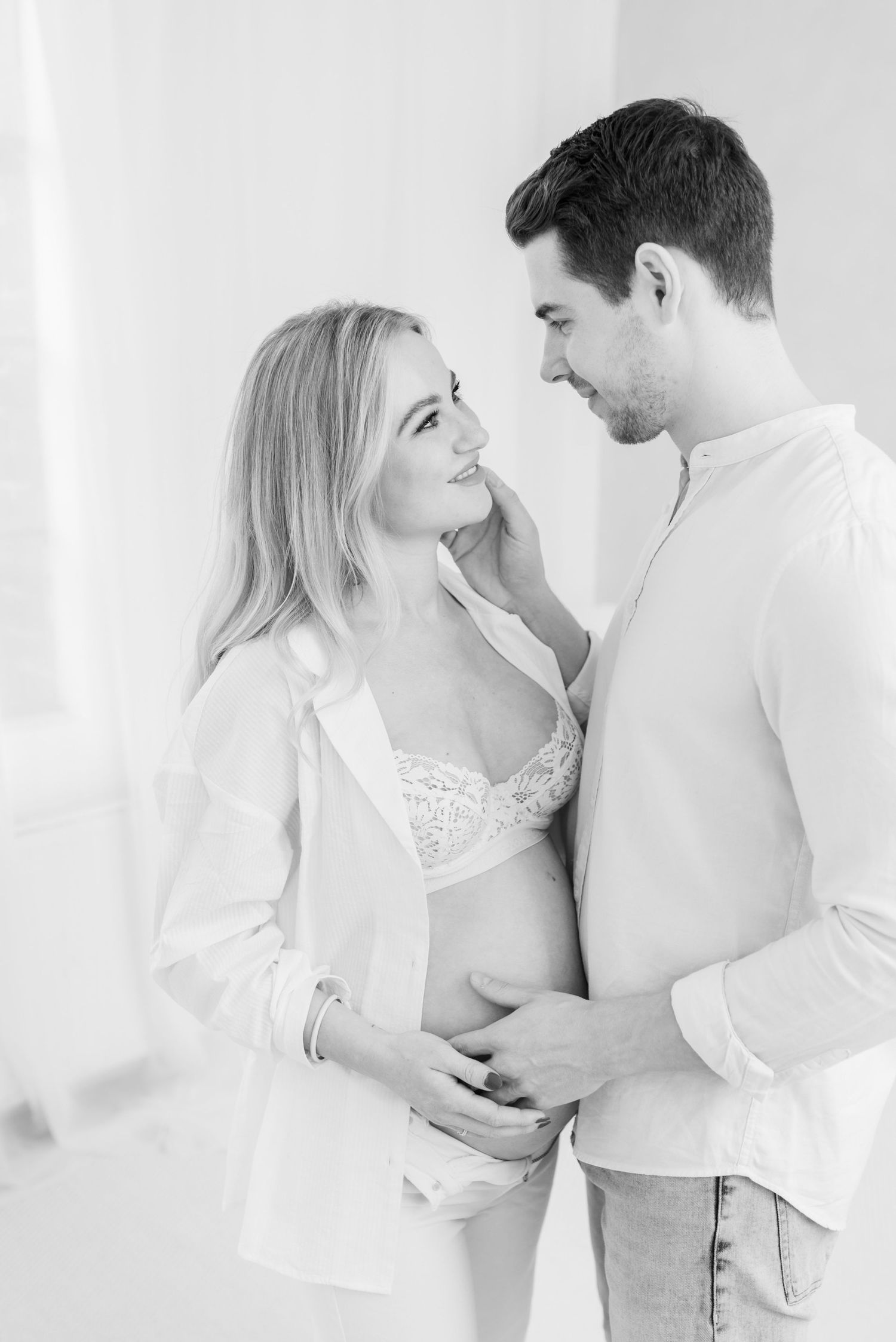 A tender black and white maternity photo showing a couple in white clothing sharing an intimate moment.