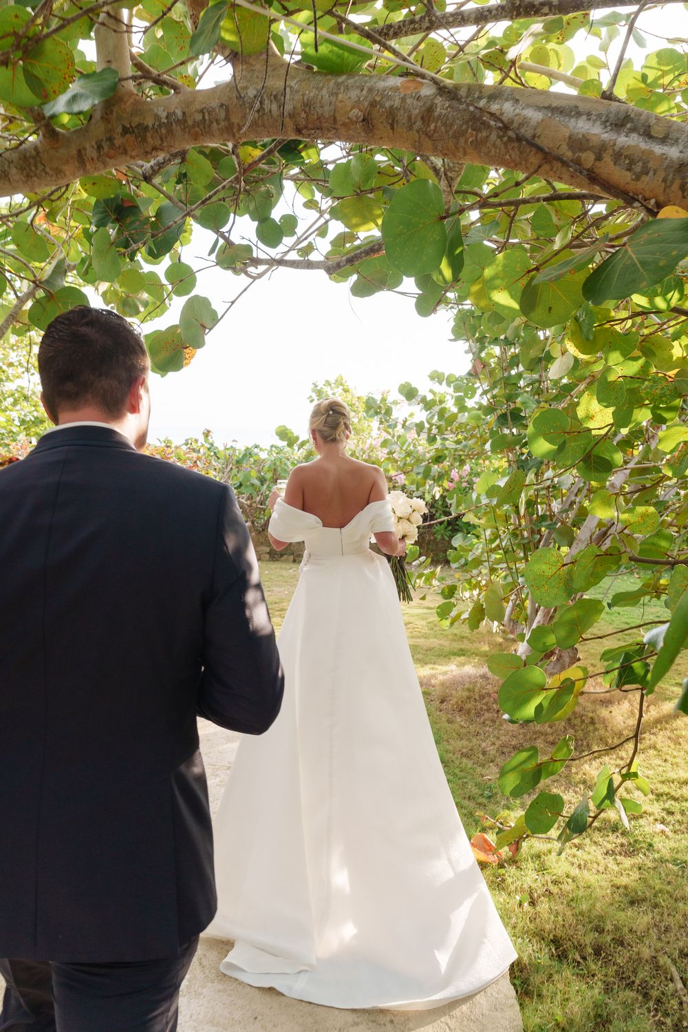 Back view of a person helping to lift a flowing white bridal gown train outdoors under leafy green trees.