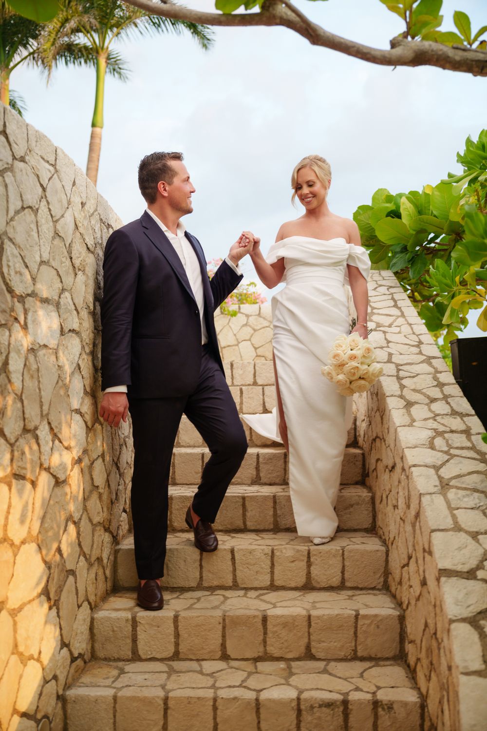 A couple in formal attire stand together on stone steps at a tropical wedding venue with palm trees and greenery.