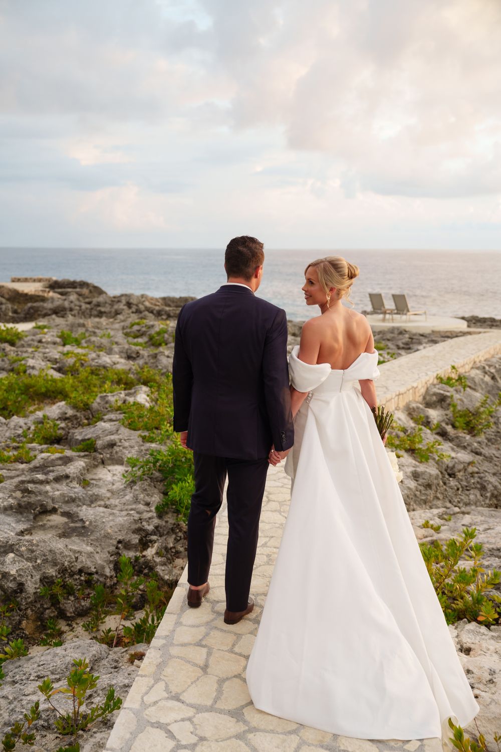 Couple stands on stone path overlooking ocean at sunset in formal wedding attire.