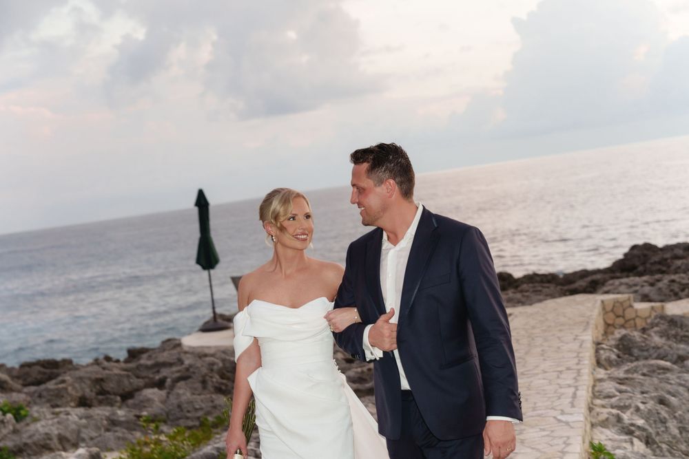 Couple shares a romantic moment walking along rocky ocean coastline at sunset in wedding attire.