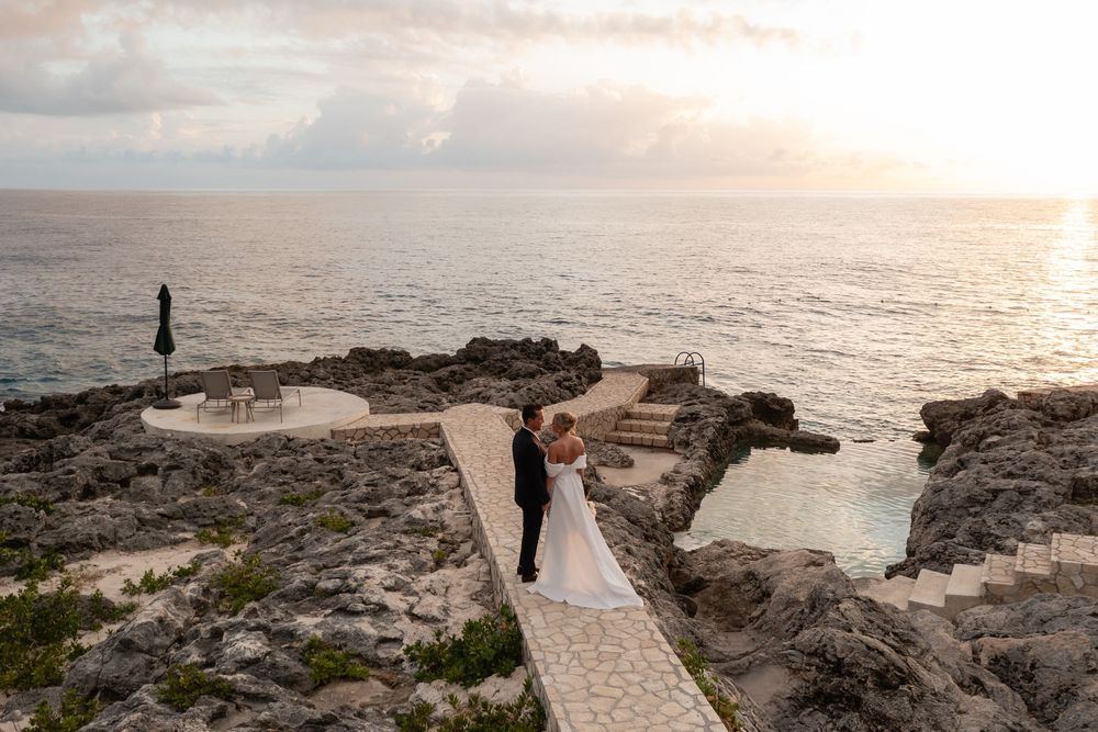 Two silhouettes stand on a rocky coastal path overlooking the ocean at sunset with dramatic clouds in the sky.