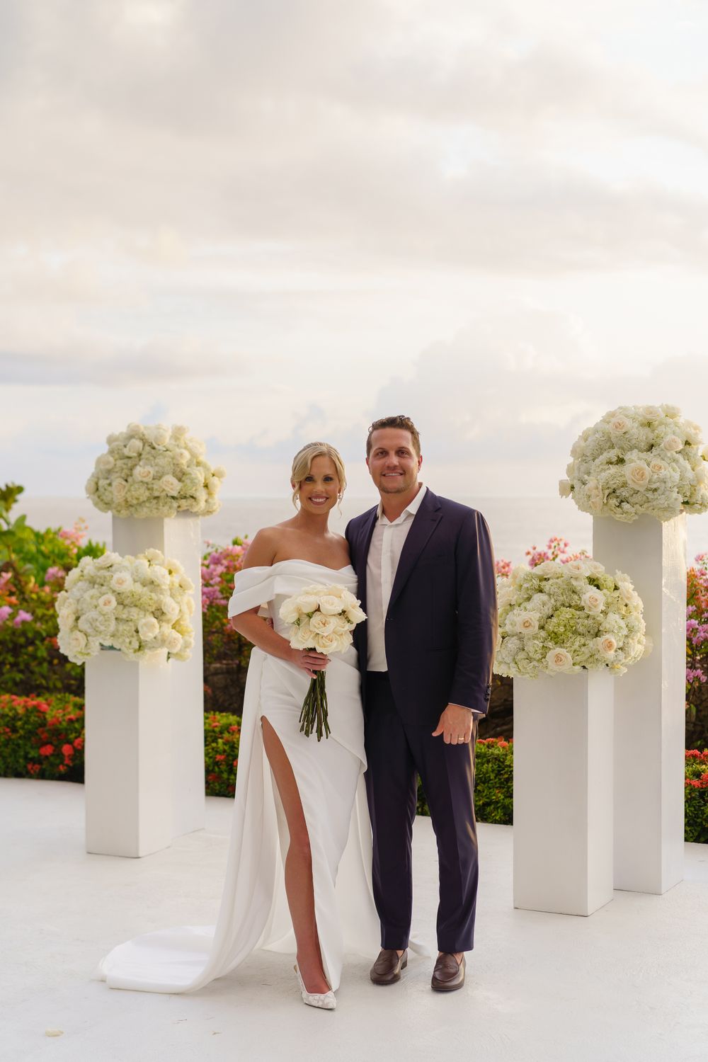 Elegant wedding ceremony setup with white floral arrangements on pedestals against a sunset sky.