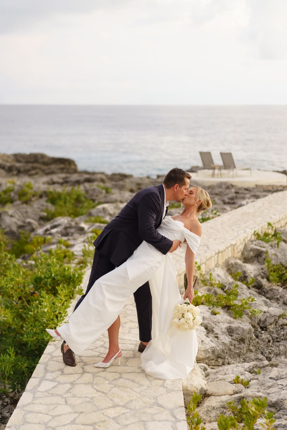 Romantic wedding couple sharing intimate moment on coastal stone pathway overlooking ocean.