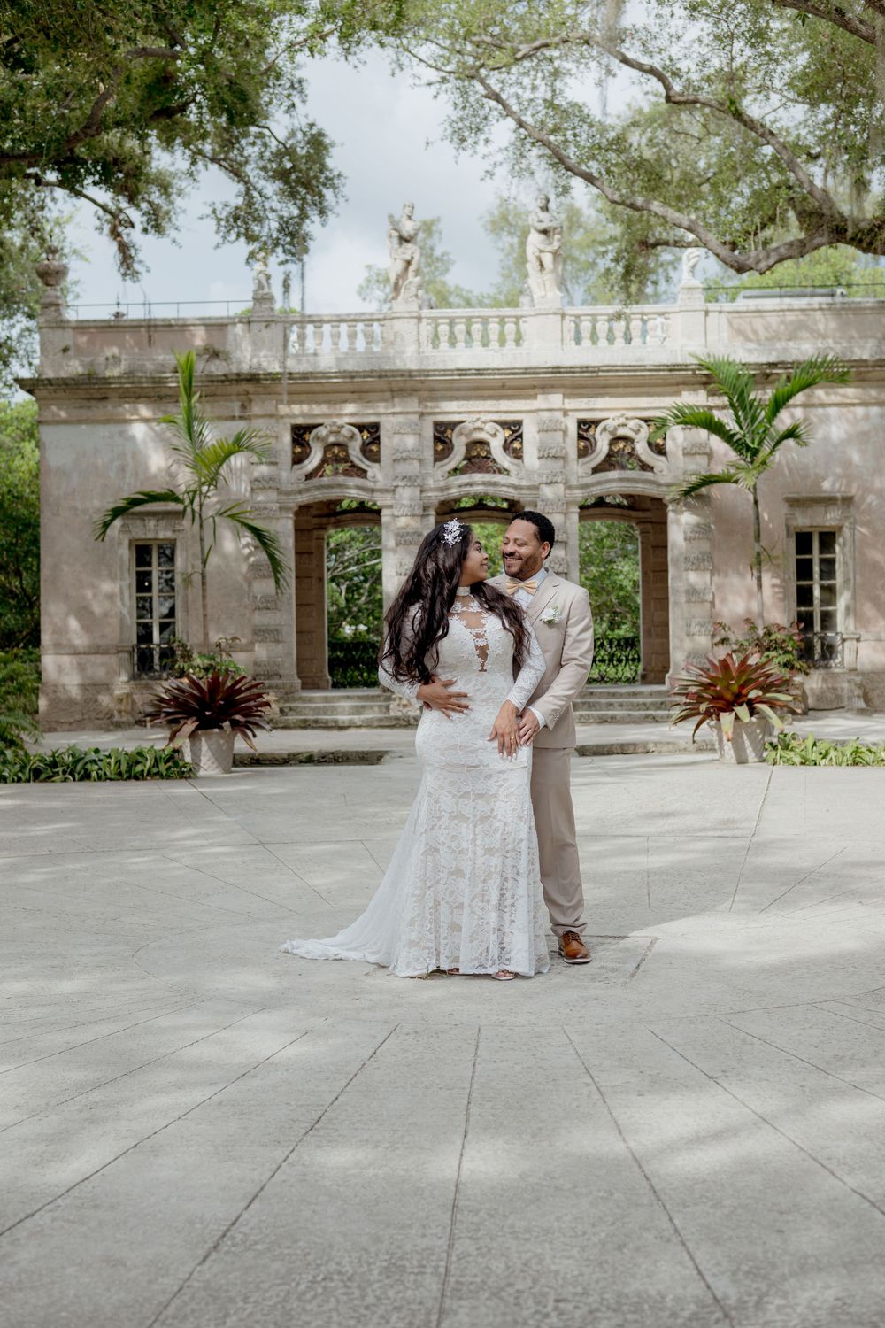 A wedding couple poses in front of an elegant stone colonnade with decorative architectural details and potted plants.
