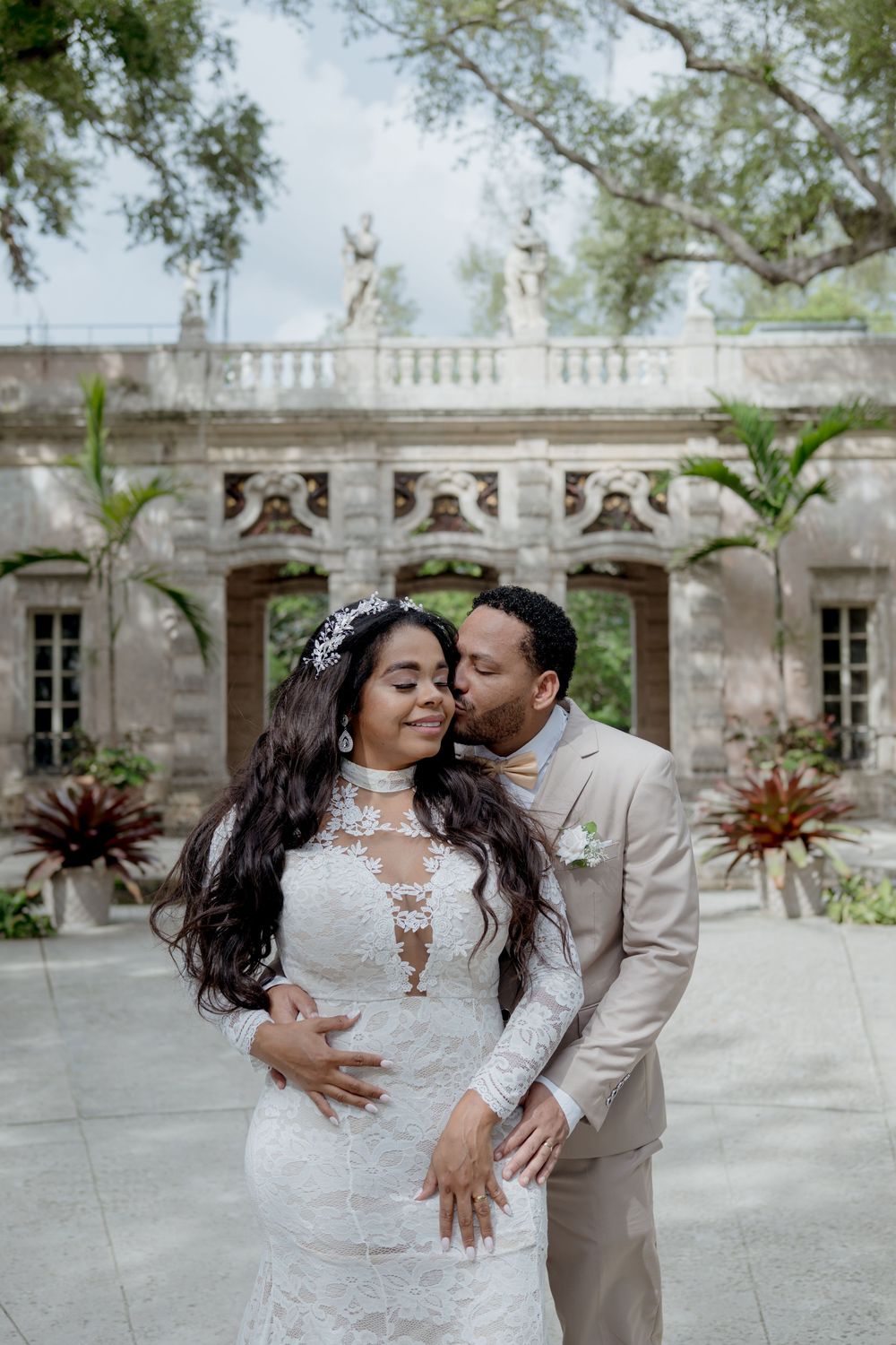 A romantic couple poses in formal wedding attire among white stone architecture and tropical plants.