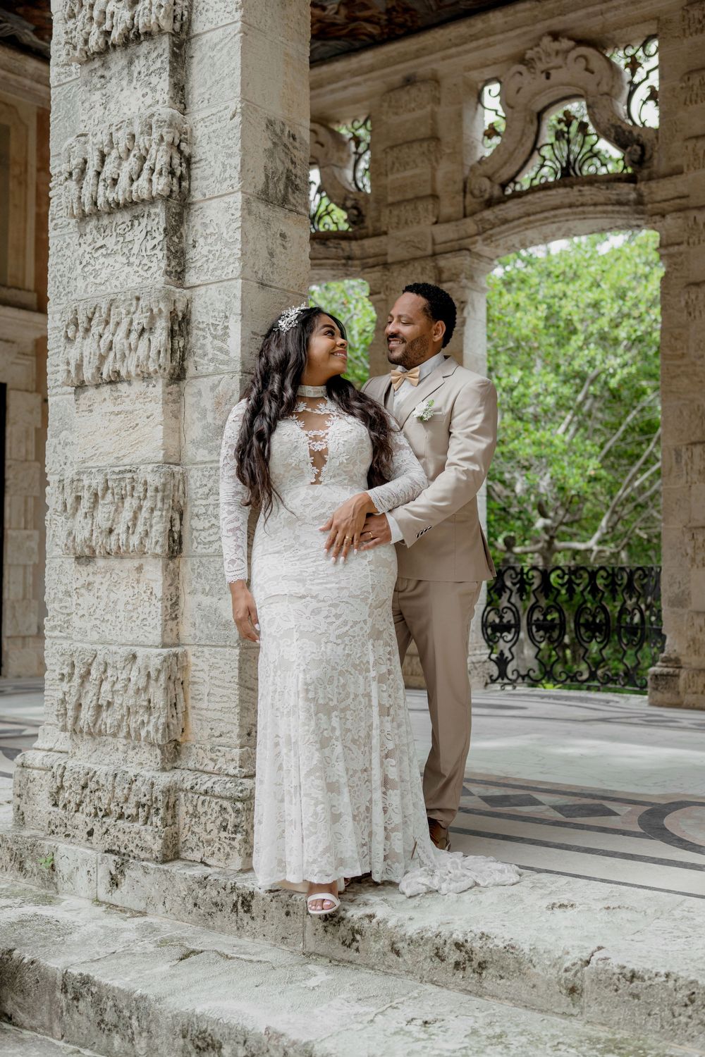 A couple shares an intimate moment in an elegant architectural setting with stone columns and ornate arches.