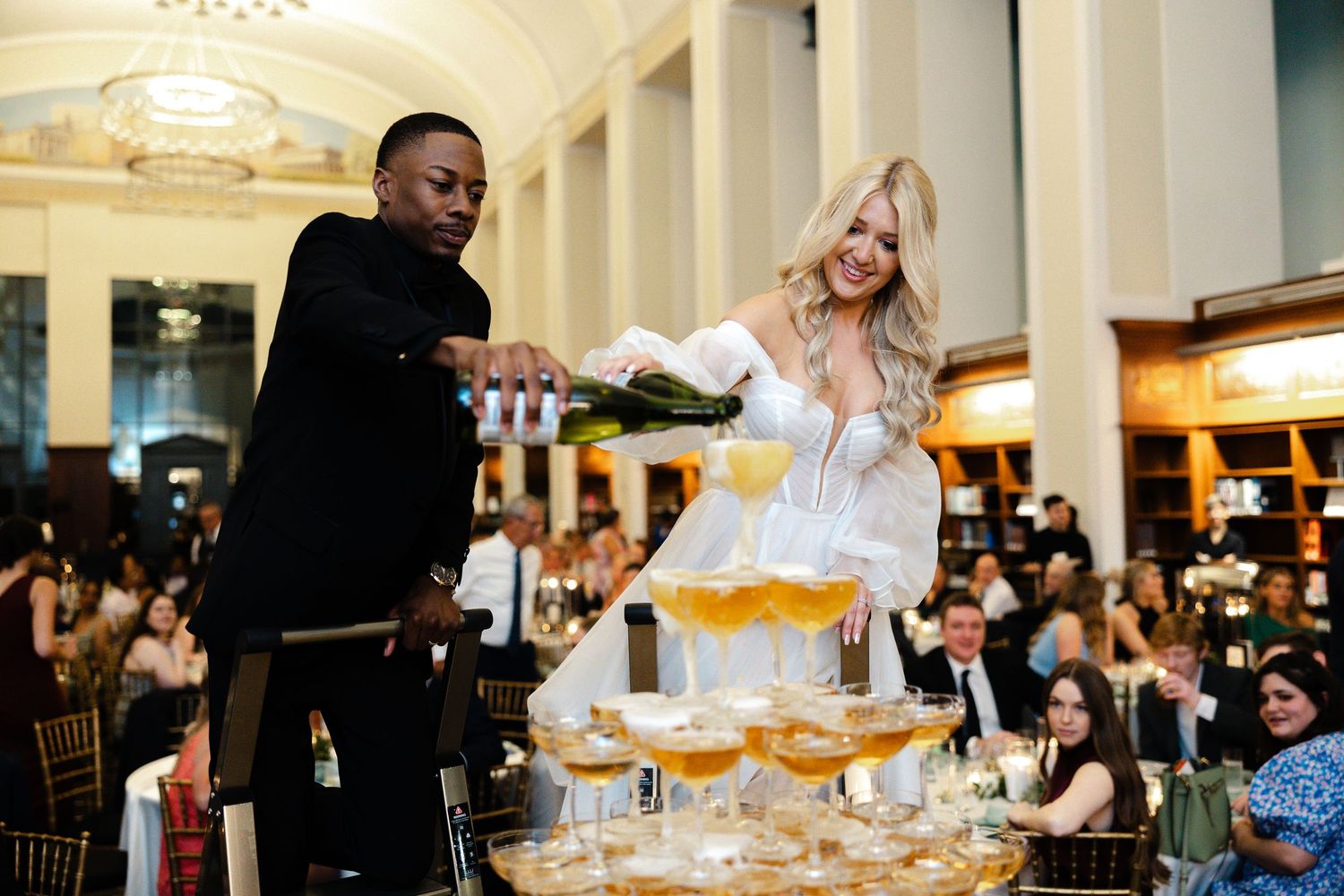 Newlyweds pour champagne into a tower of glasses at their reception celebration in an elegant indoor venue.