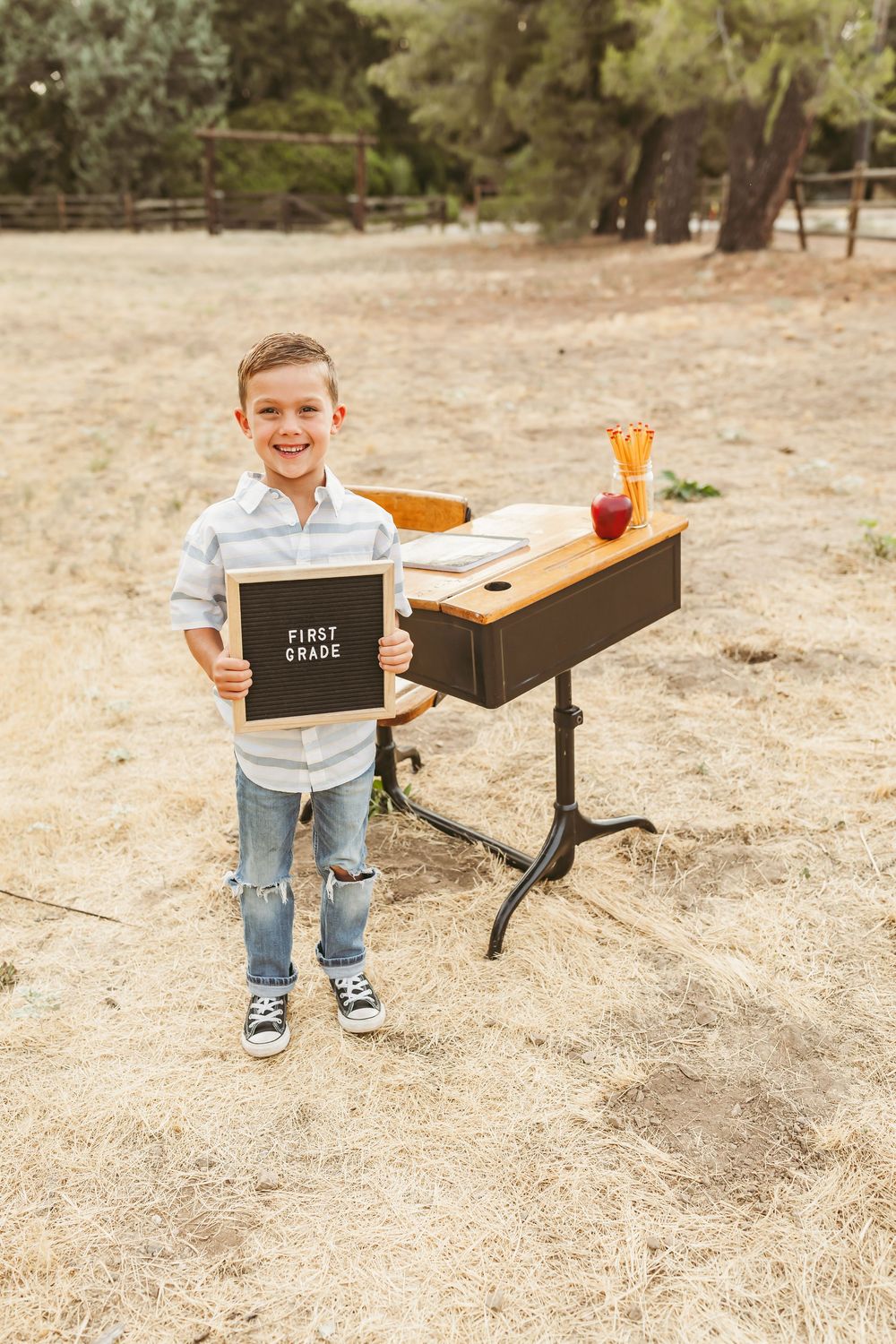Student stands outdoors holding a first grade sign near a vintage school desk in a park setting.