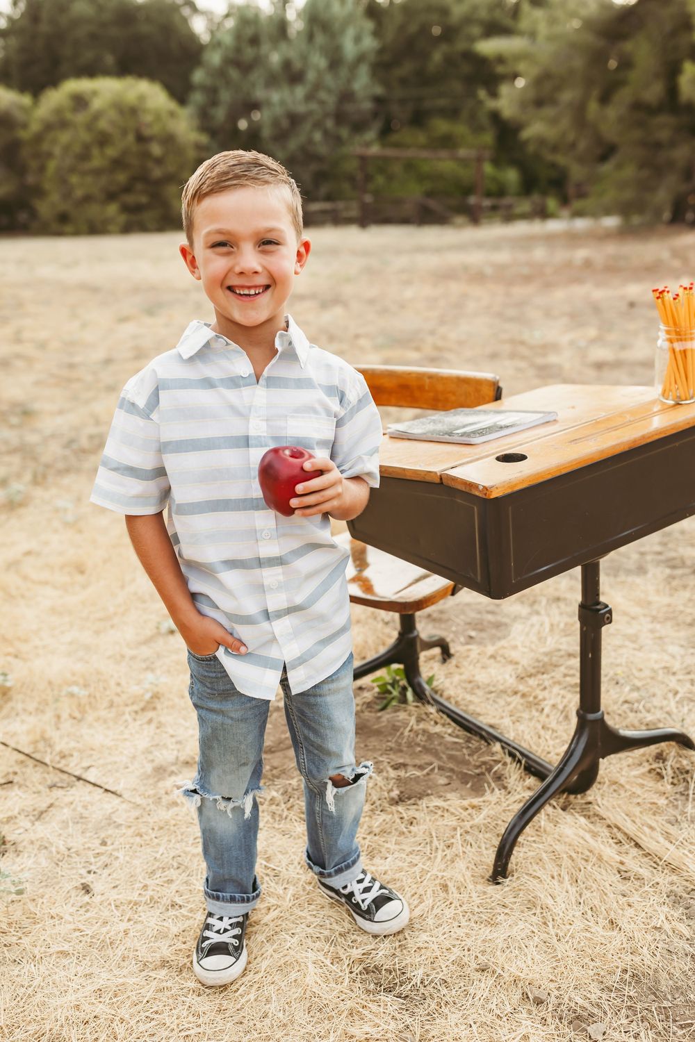 Smiling child in striped shirt and jeans holds red apple while standing next to outdoor table on dry grass.
