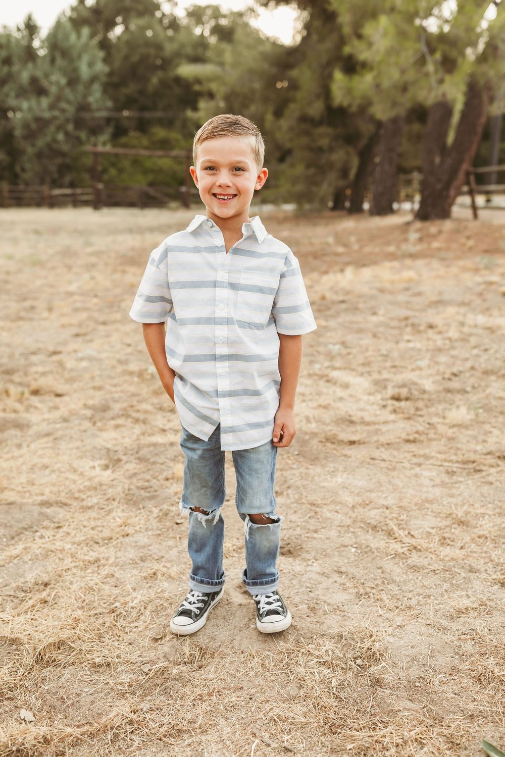 Young student in striped shirt and ripped jeans stands smiling in an outdoor school setting.