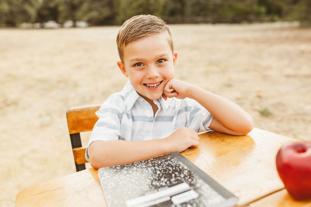 A cheerful student sits at a wooden desk with school supplies and a red apple nearby.