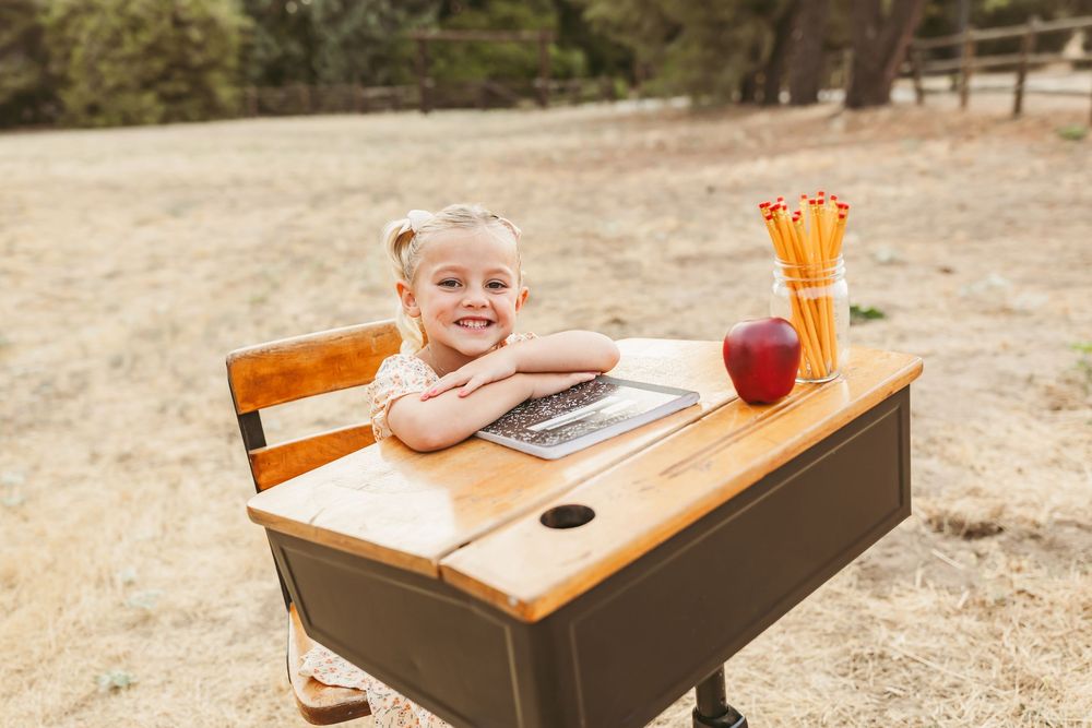 Student sits at a vintage wooden school desk outdoors with supplies and a red apple on the desktop.