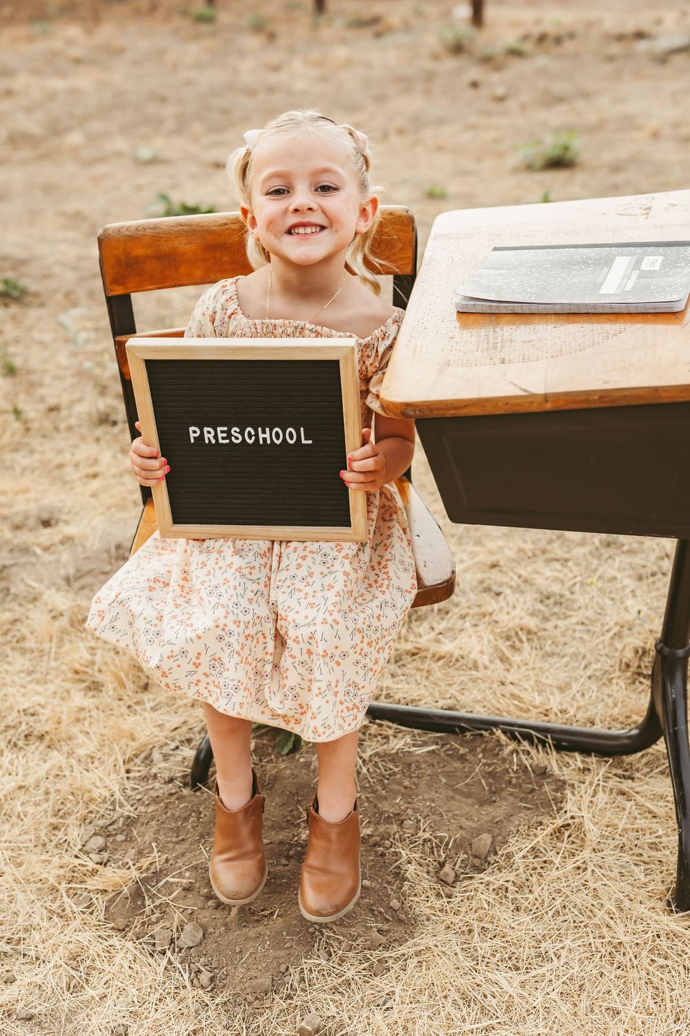 Child holding preschool sign board while sitting at vintage wooden school desk outdoors on brown ground.