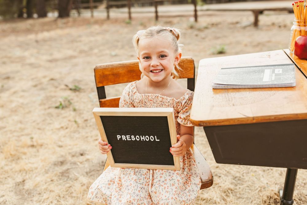 Student in dress holds preschool sign while standing by wooden desk outdoors during back-to-school photoshoot.