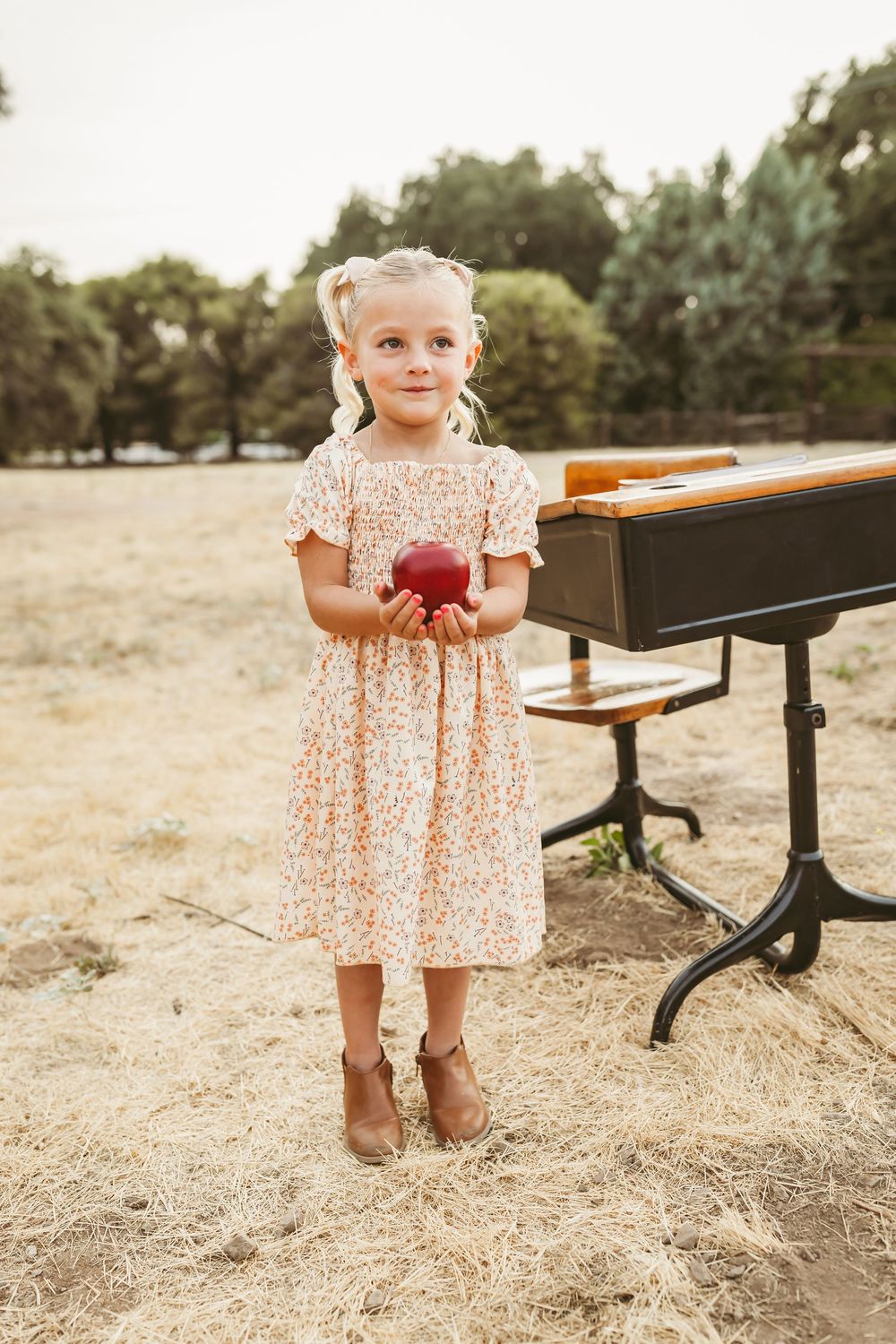 A small child in a floral dress stands holding an apple next to a piano outdoors at sunset.