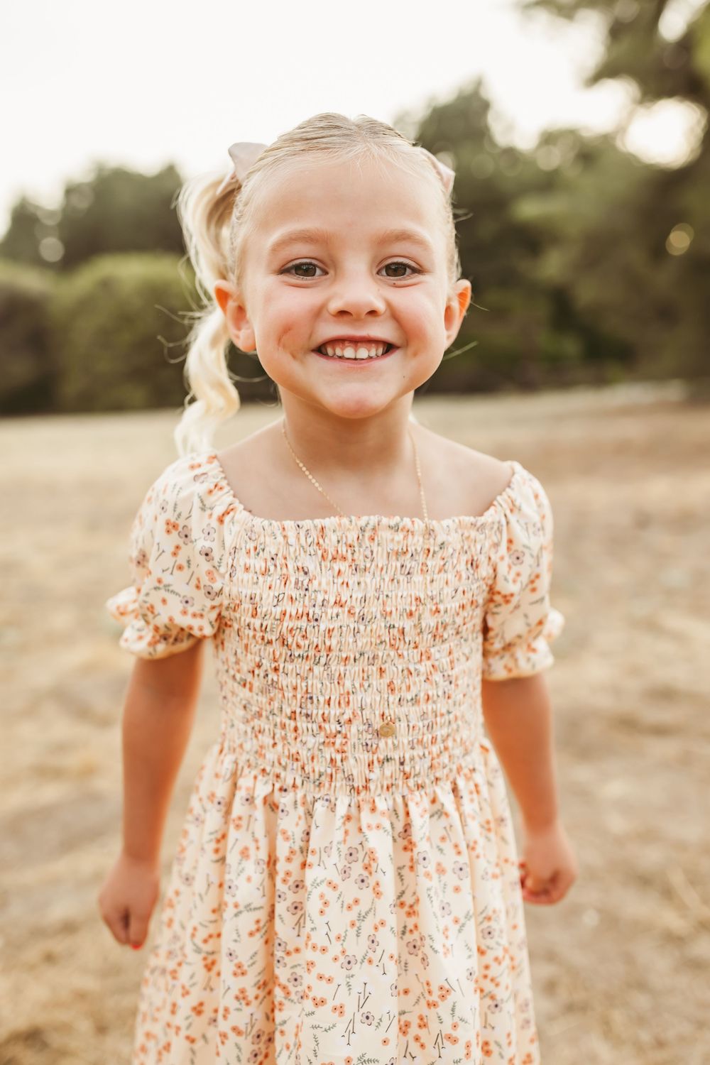 Young child wearing a floral smocked dress smiles brightly in warm evening light.