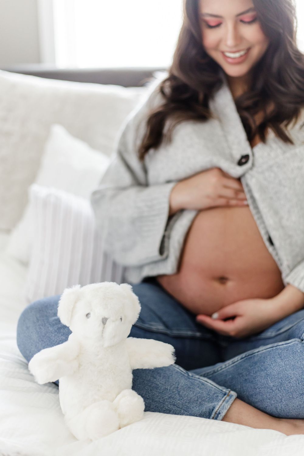 A pregnant woman in jeans and gray sweater reclines on white bedding with a small white teddy bear.