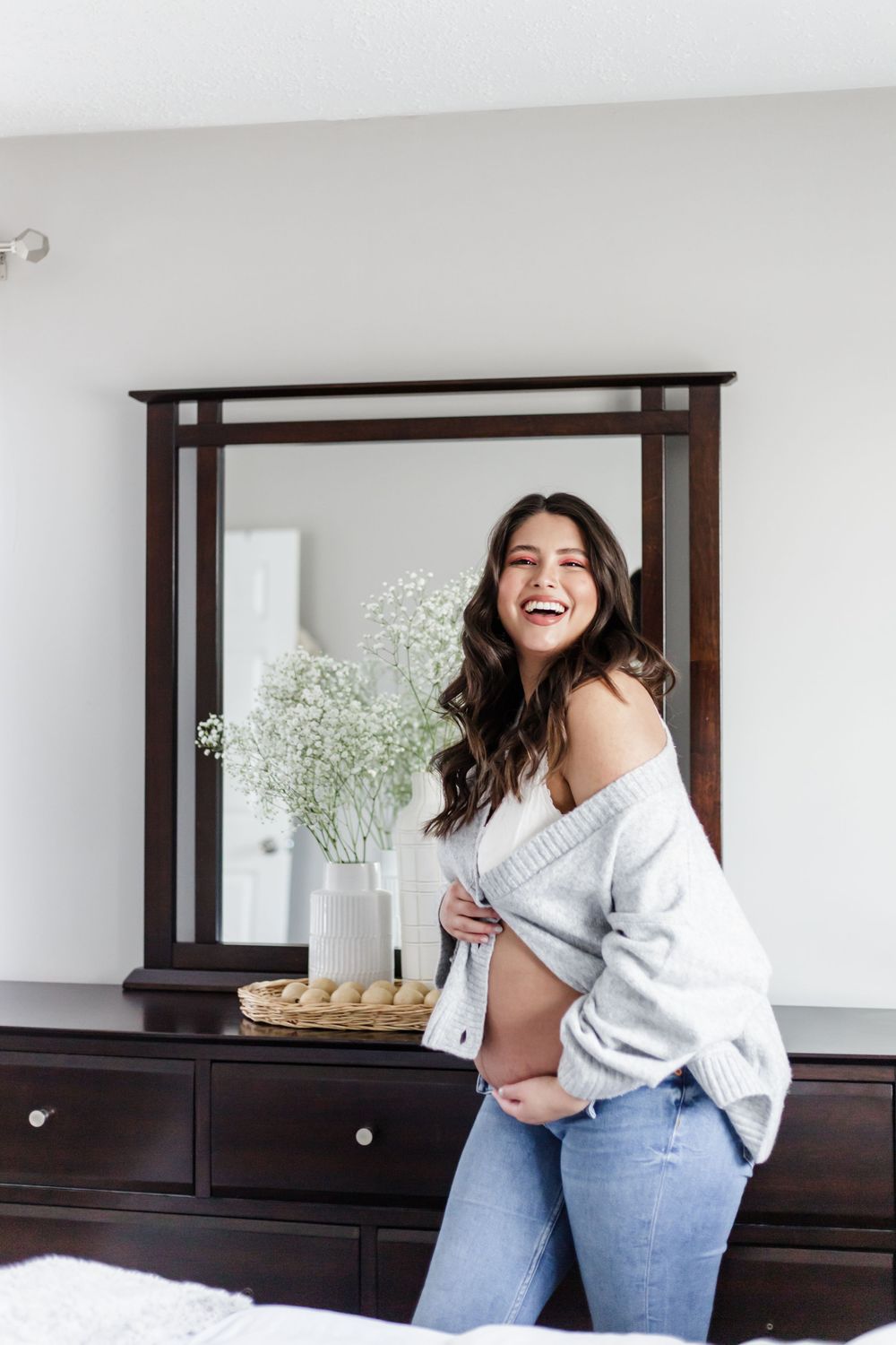 Woman in off-shoulder sweater laughs joyfully near dresser with mirror and baby's breath flowers.
