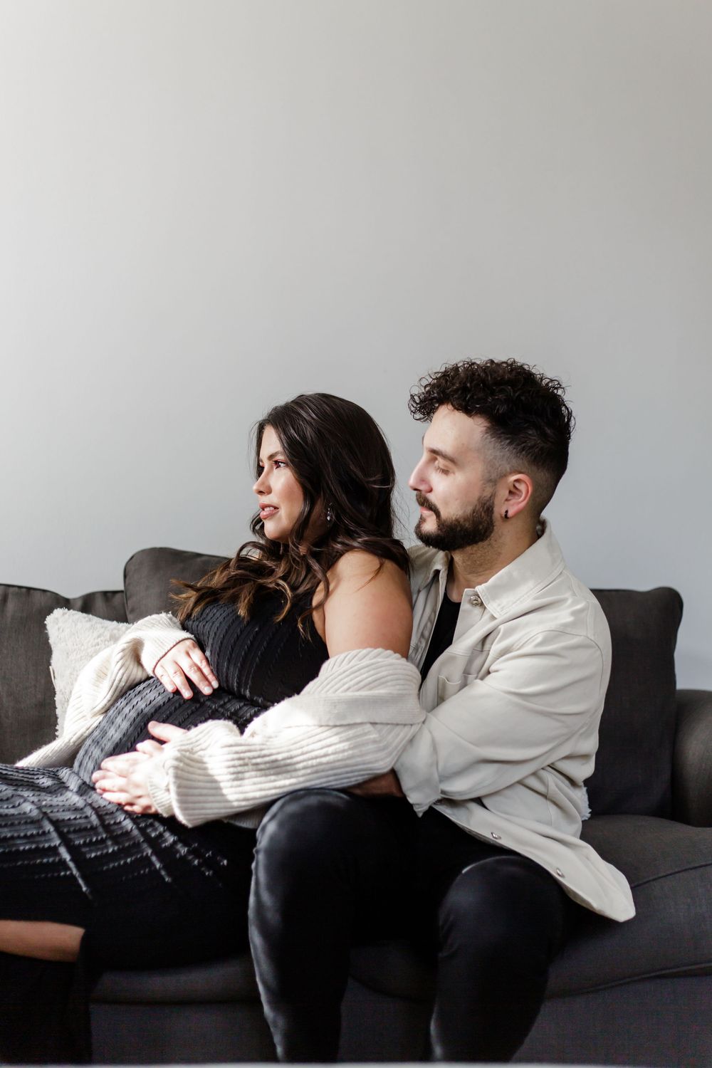 A couple lounging together on a dark couch in casual white shirts and dark pants.