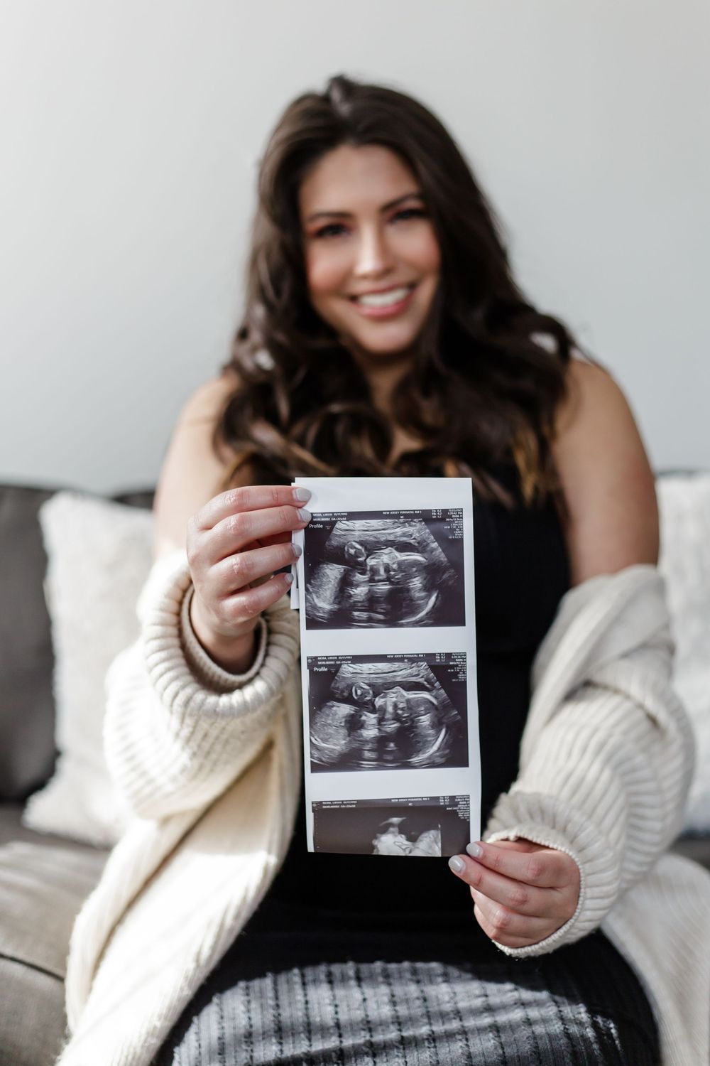Woman in white sweater holds ultrasound photos while sitting at home with joyful pregnancy announcement expression.