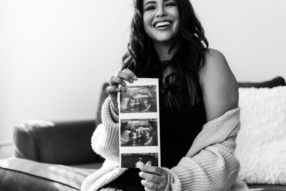 Black and white photo showing someone sitting on couch holding ultrasound photos while laughing joyfully.