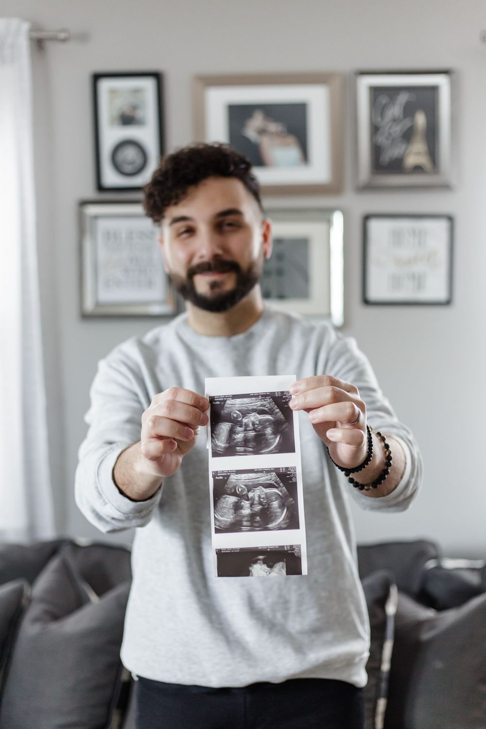 Someone in a gray sweatshirt holds up ultrasound photos while standing in front of a gallery wall with framed artwork.