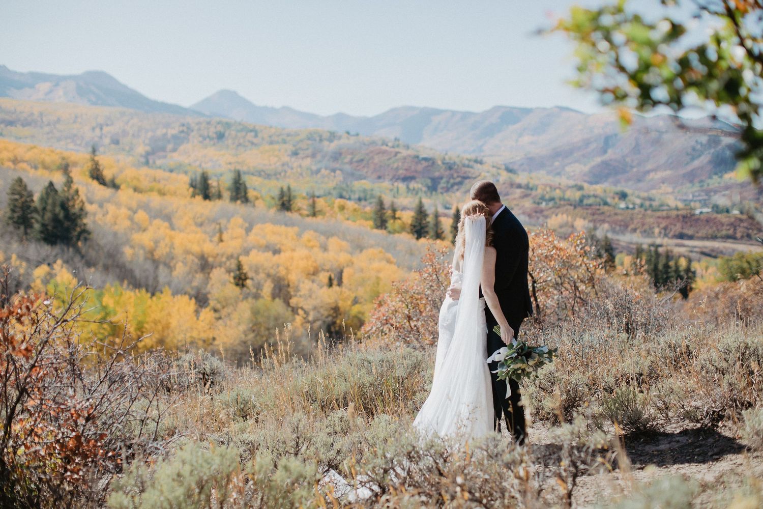 Couple embracing while standing in a fall mountain landscape with golden foliage and distant peaks. Aspen elopement photography.