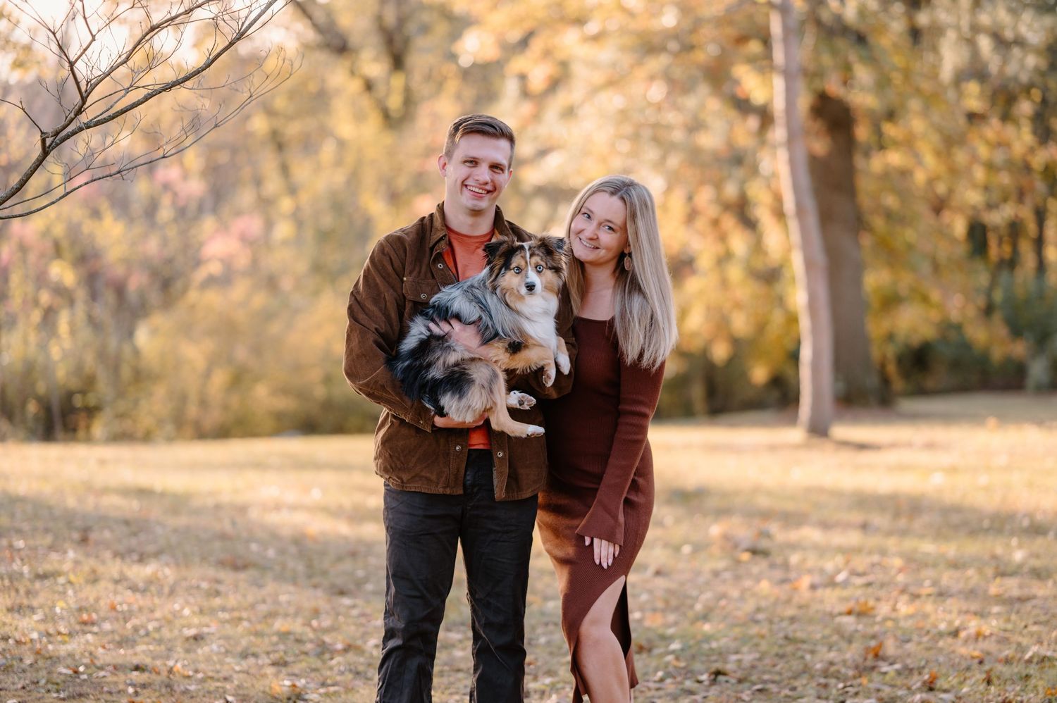 People in rustic fall outfits with their dog pose outdoors for photos in a park during golden hour.