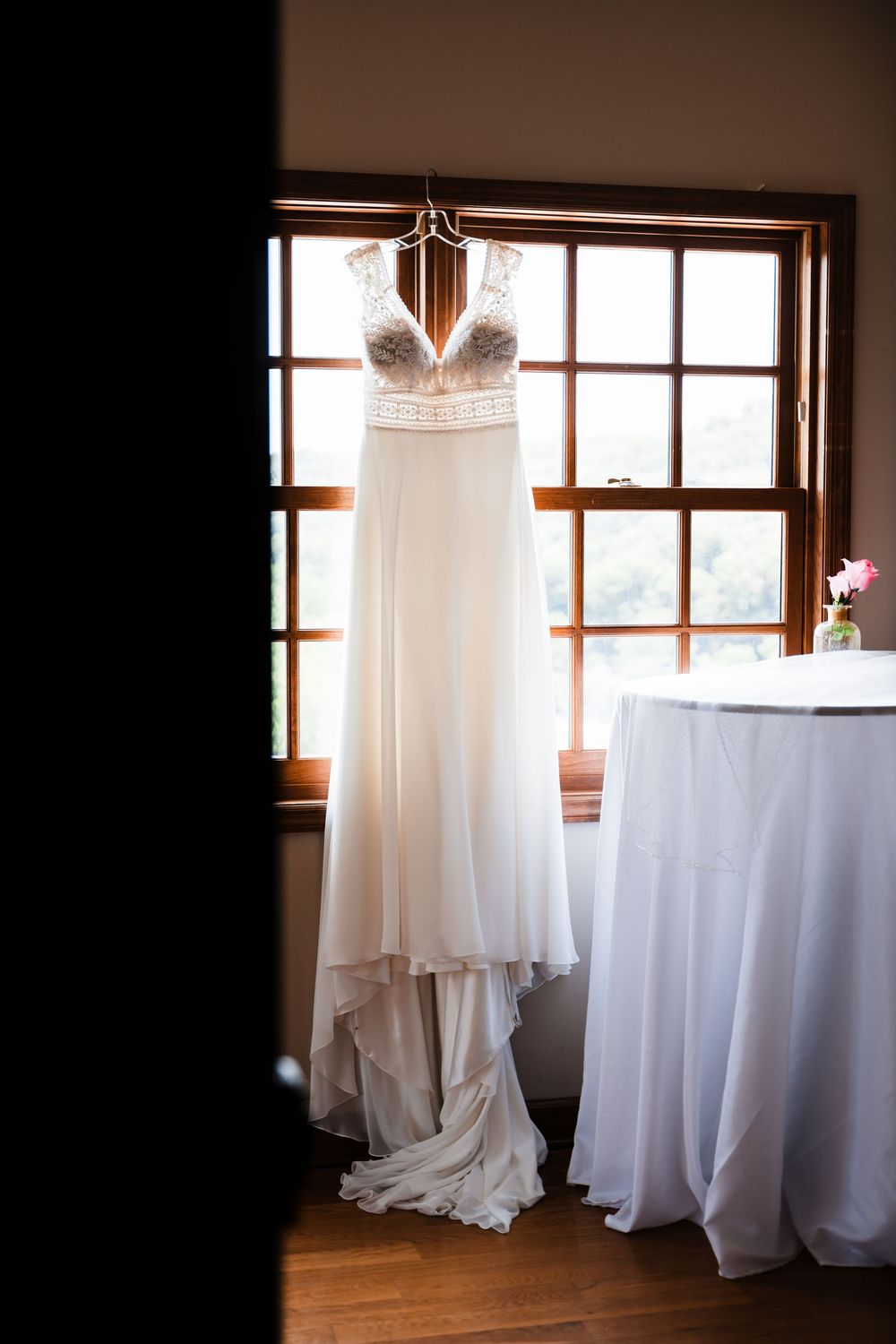 White wedding dress hanging by windows in a rustic indoor setting with natural light.