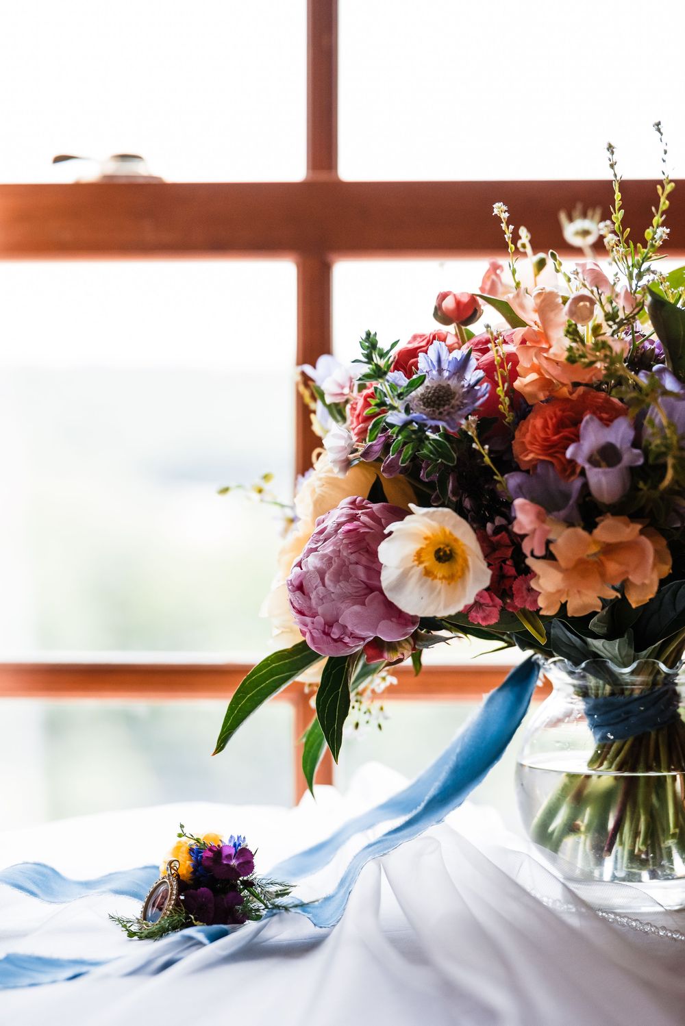 Elegant floral arrangement with peonies and mixed flowers against window light with blue ribbon accent.