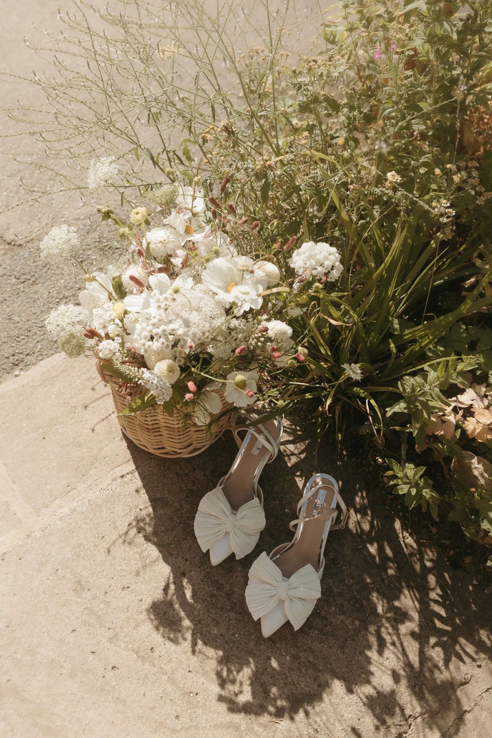 Elegant white bow wedding shoes next to woven basket filled with delicate white floral arrangements in sunlight.