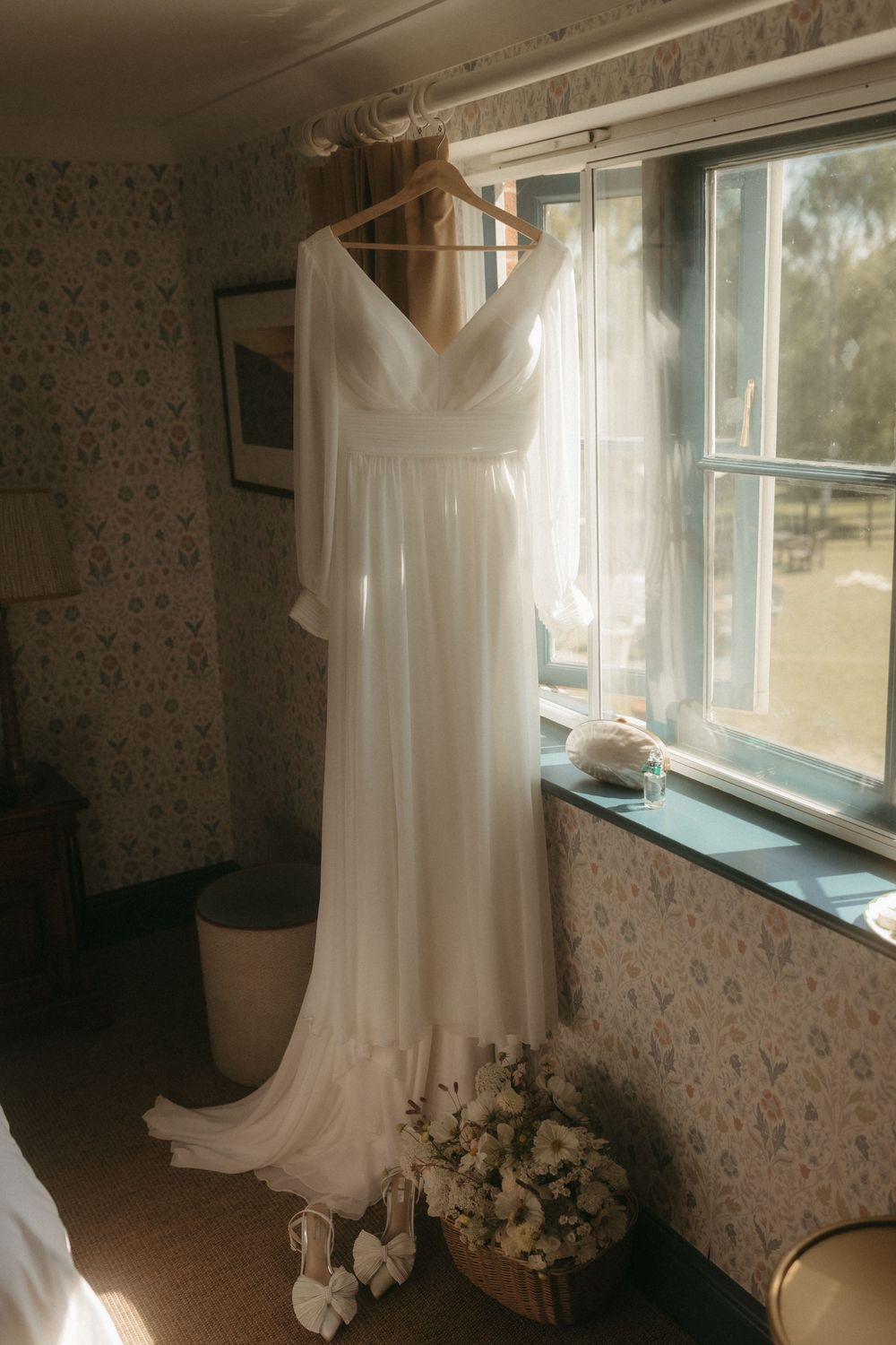 A flowing white wedding dress hangs elegantly by a sunlit window with floral wallpaper and fresh bouquets beneath it.