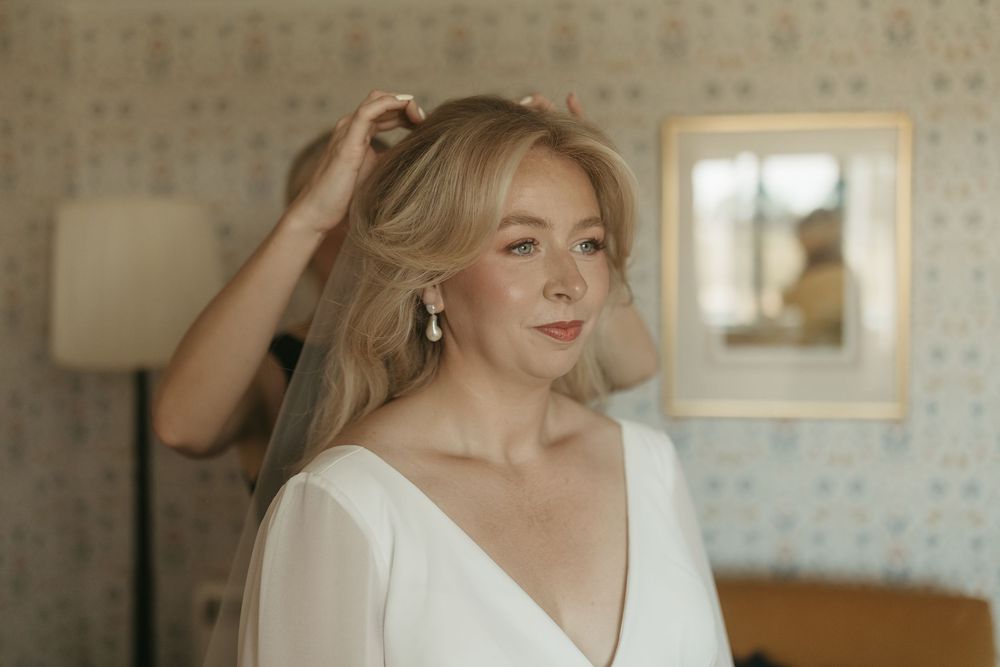 A person in a white dress gazes thoughtfully while standing in a room with floral wallpaper and natural lighting.
