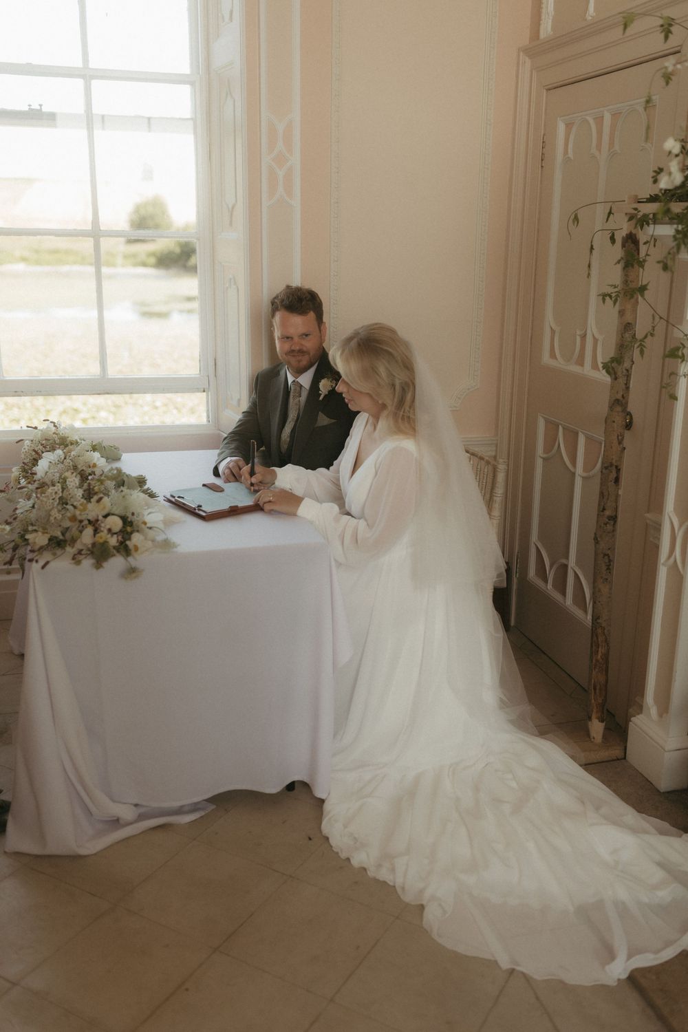 A couple signs marriage documents at a white-covered table by a window, with the bride wearing a flowing white gown and veil.